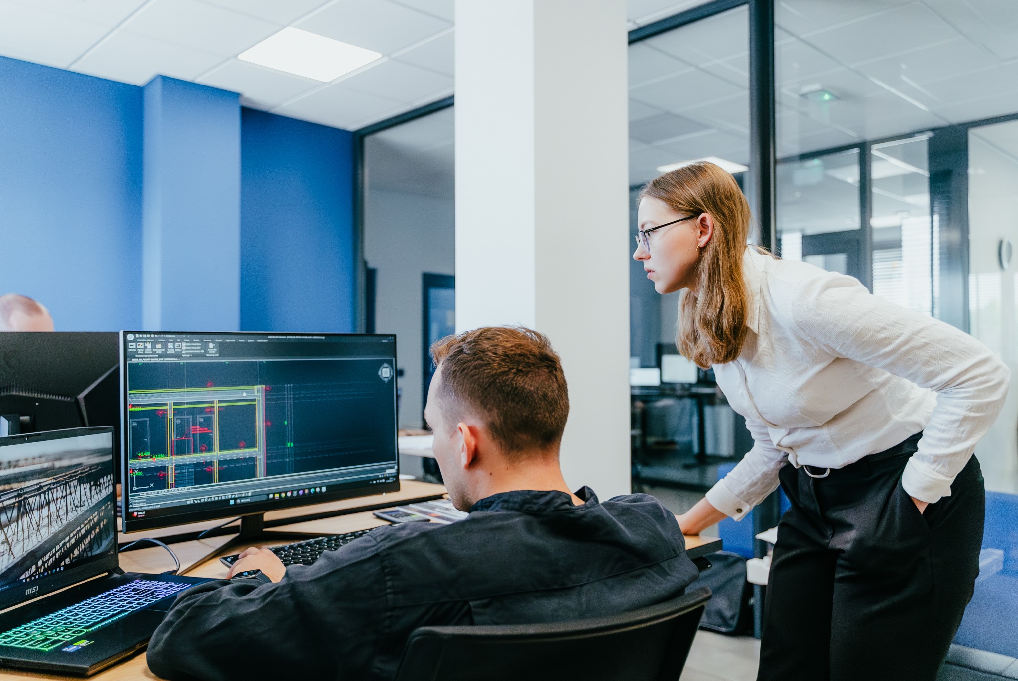 Architektura wnętrz biura: mężczyzna pracuje przy komputerze z widocznym projektem budowlanym, kobieta w okularach pochyla się, obserwując ekran.