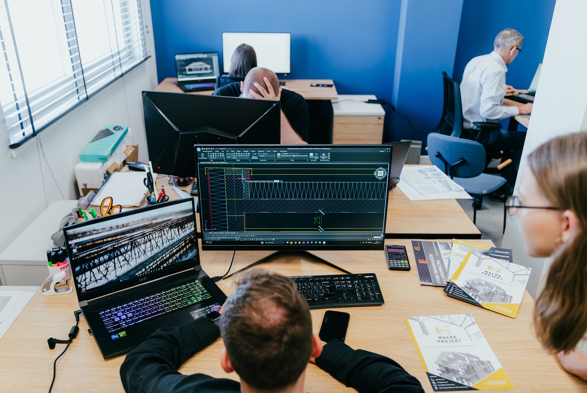 Biuro projektowe w Radzionkowie: widok zza pleców projektanta pracującego nad projektem konstrukcyjnym na ekranie komputera, obok laptop z otwartym programem graficznym, w tle inni pracownicy przy...