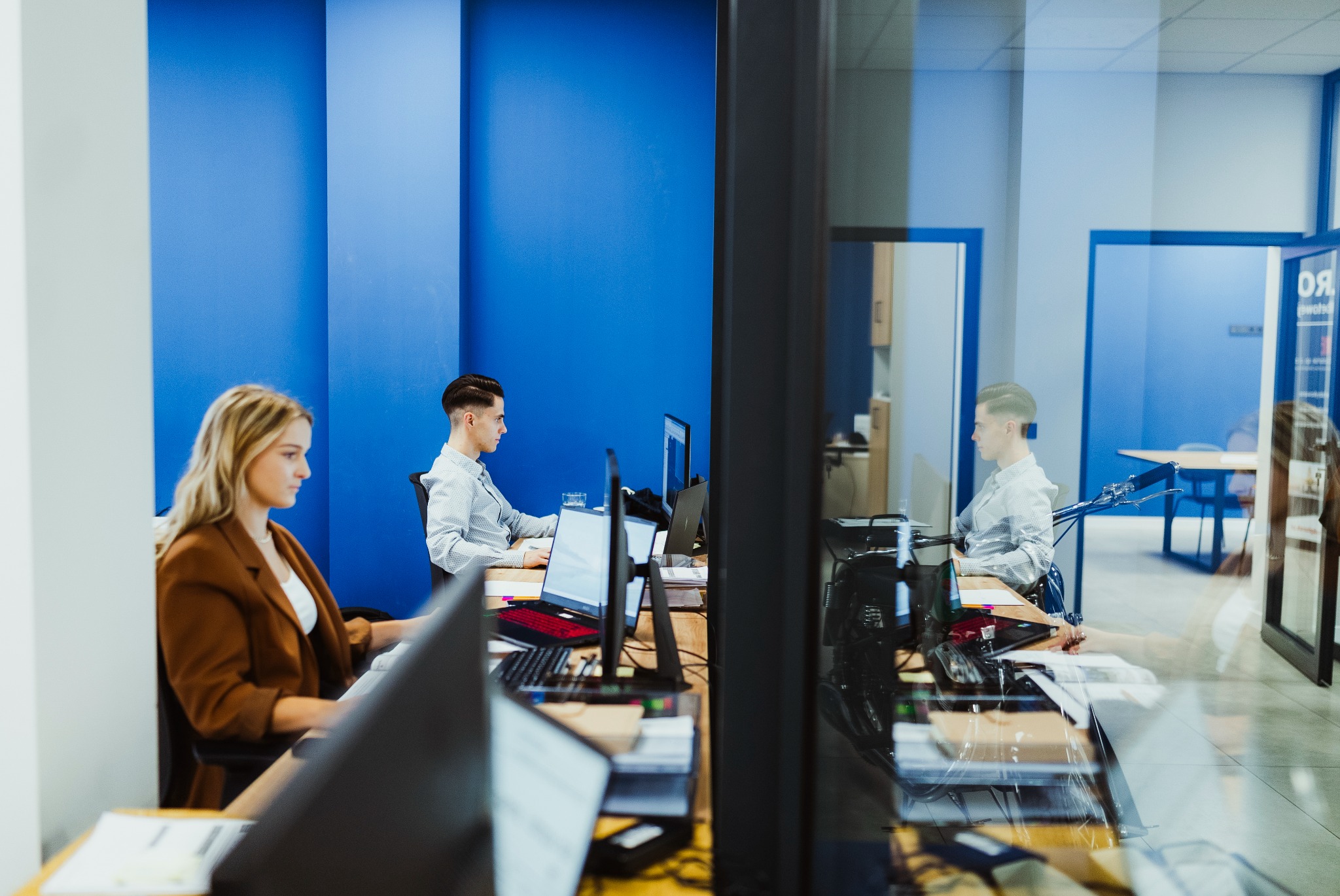 Wnętrze biura z dwoma pracownikami przy biurkach, na których stoją monitory i laptopy z czerwonymi klawiaturami, niebieskie panele ścienne w tle.