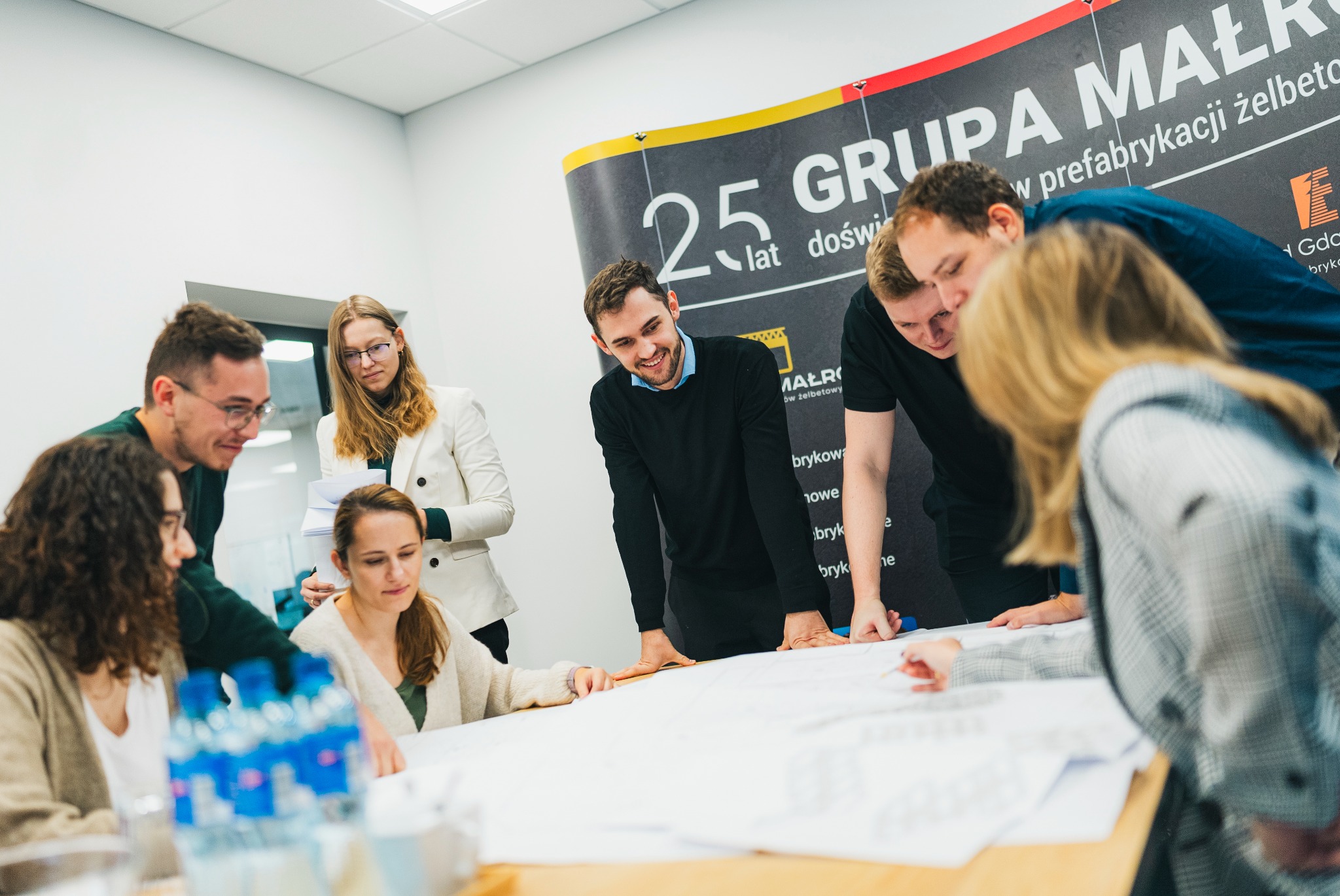 Zespół inżynierów i architektów analizuje plan budynku na dużym stole w jasnym biurze, w tle widoczny baner reklamowy firmy budowlanej.