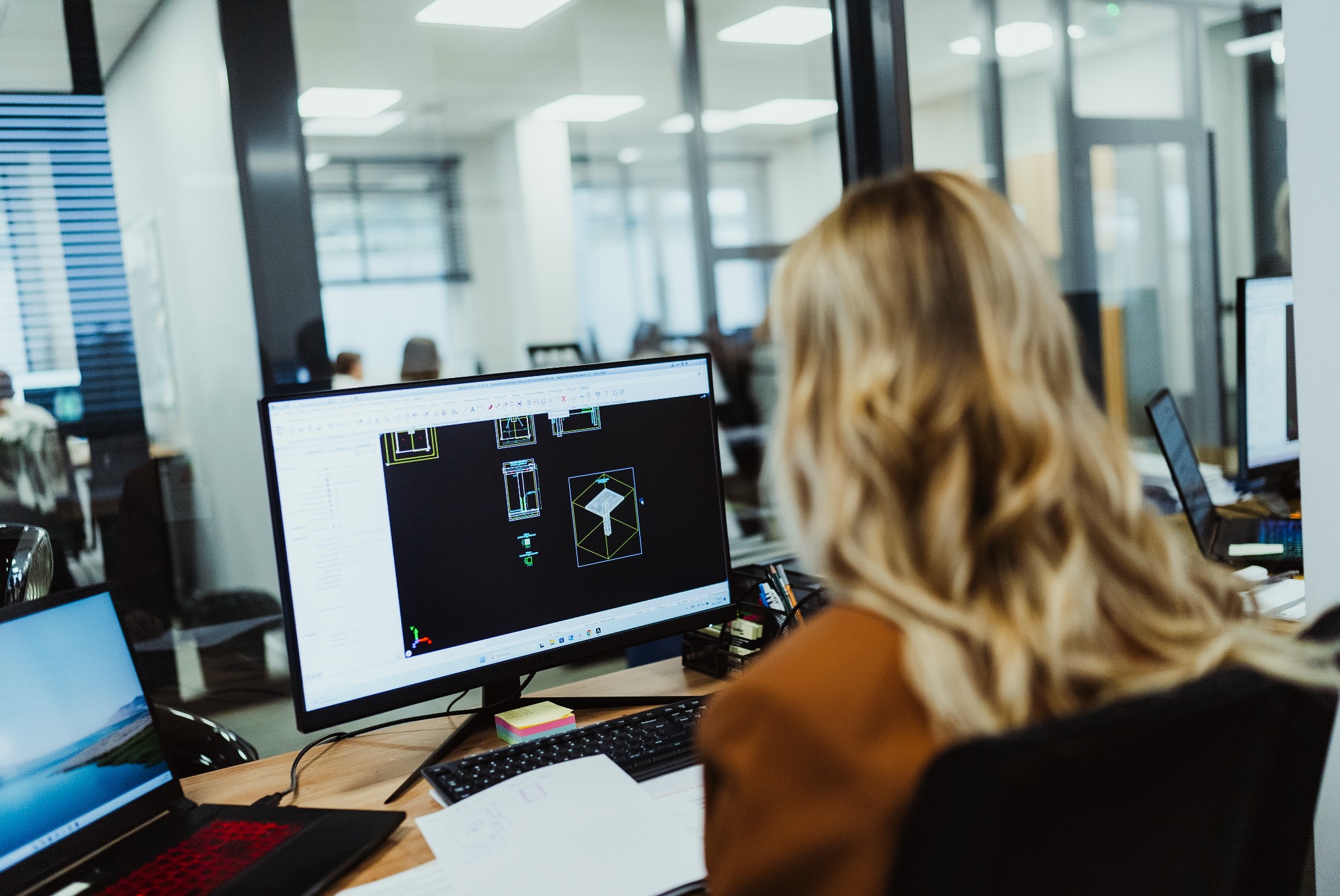 Kobieta z blond włosami siedzi przy biurku w biurze, patrząc na monitor komputera wyświetlający schematyczny projekt architektoniczny. Na biurku widoczny laptop, klawiatura, notatki i inne...