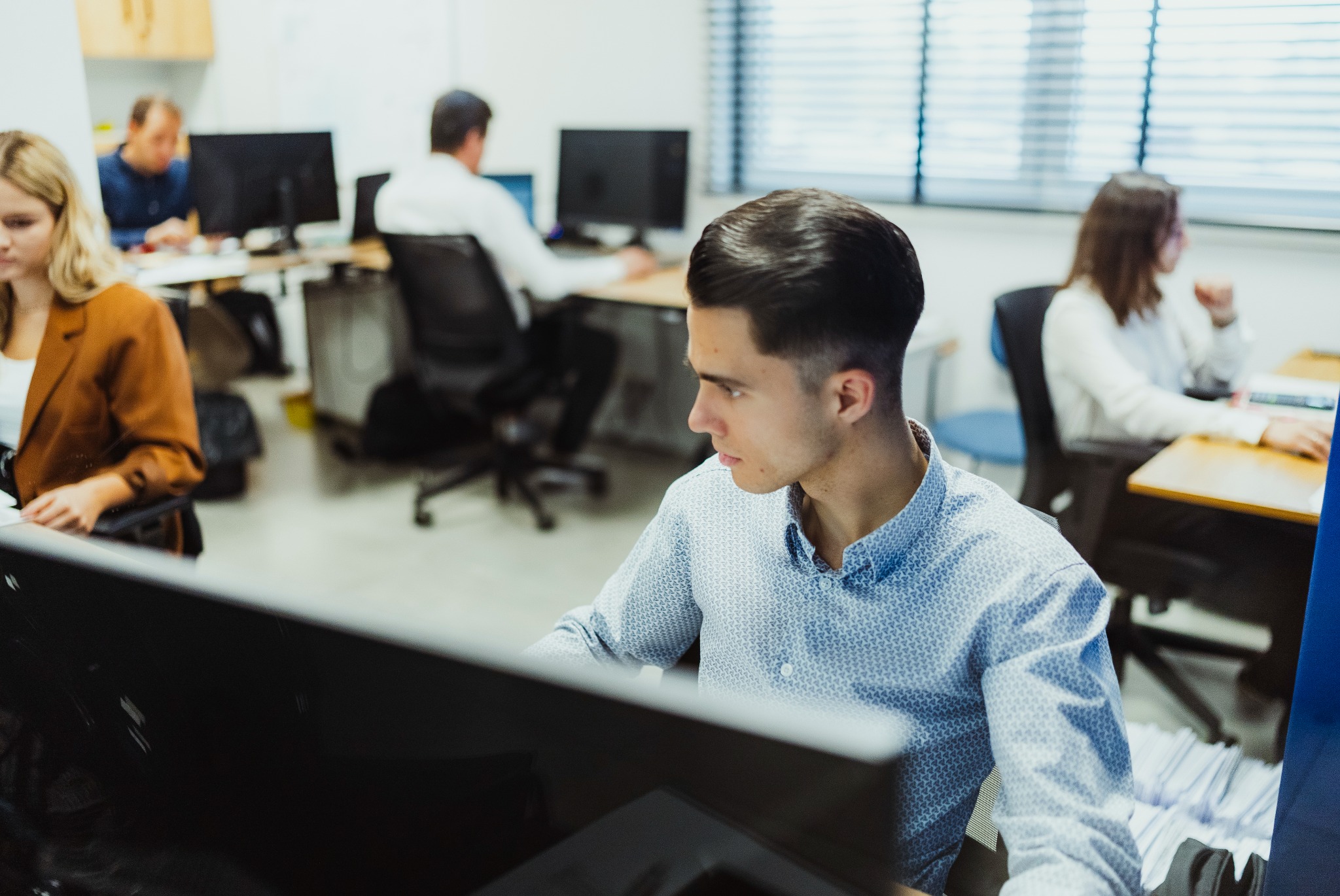 Młody mężczyzna w błękitnej koszuli skupiony na pracy przy komputerze w nowoczesnym biurze z innymi pracownikami w tle.