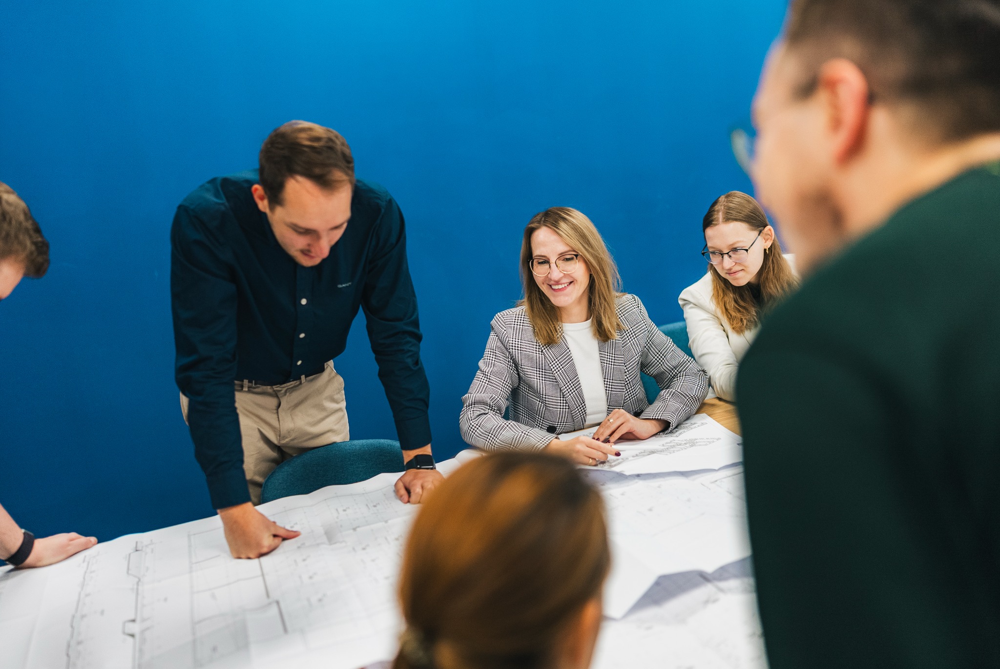 Zespół architektów analizuje plany budynku na dużym stole w biurze z niebieską ścianą, kobieta w okularach uśmiecha się wskazując na szczegóły na planie.