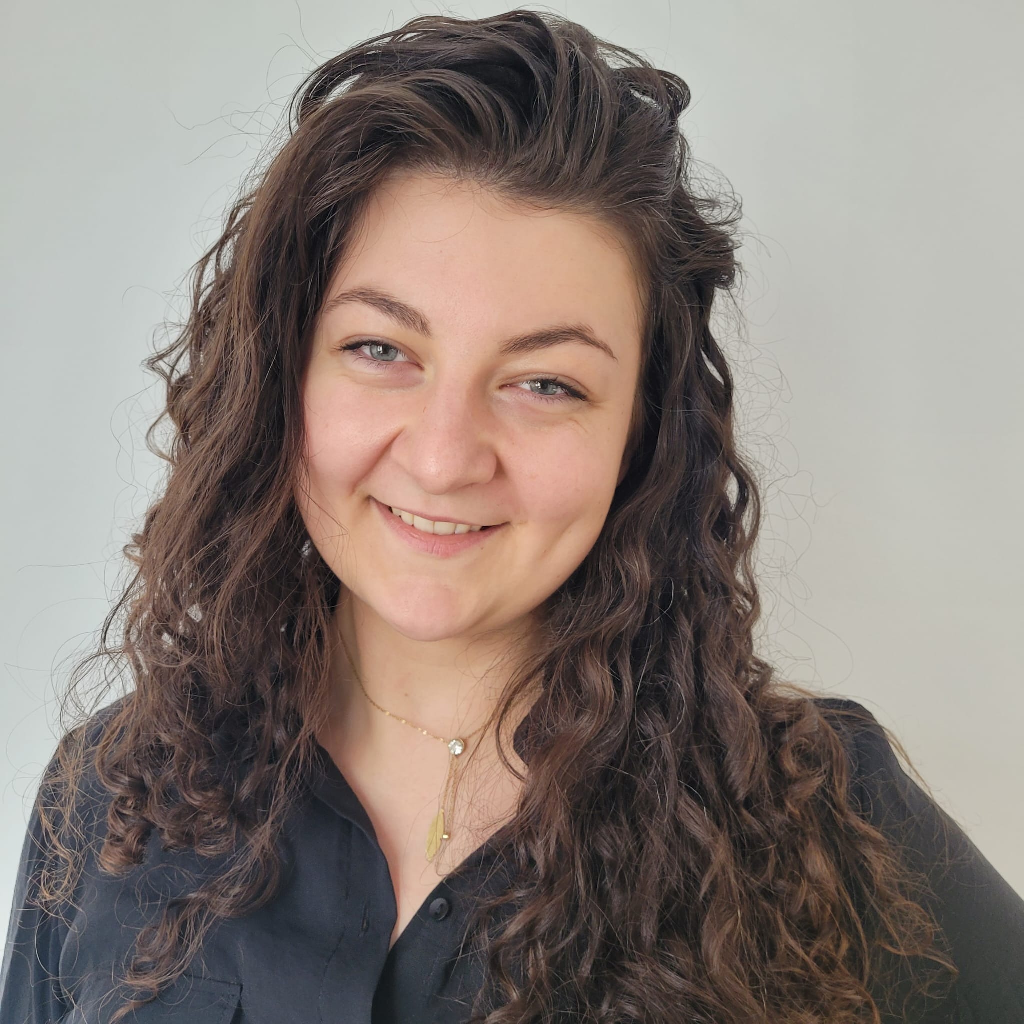 Portret uśmiechniętej kobiety z kręconymi włosami i delikatnym naszyjnikiem na tle jasnej ściany w Gryfinie.