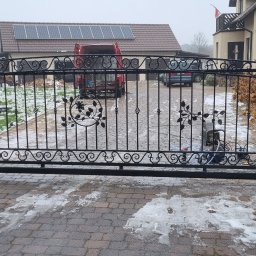 Daniel Jurkiewicz - Czarna, kuta brama wjazdowa z dekoracyjnymi elementami roślinnymi, osadzona na murowanych słupkach. W tle dom z panelami słonecznymi i częściowo ośnieżony teren.