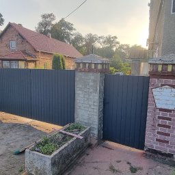 Daniel Jurkiewicz - Nowoczesne, grafitowe ogrodzenie panelowe z bramą, osadzone między murowanymi słupkami. W tle dom z czerwonej cegły i zielona roślinność.