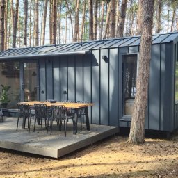 Daniel Jurkiewicz - Nowoczesny, szary domek mobilny z metalowym wykończeniem, otoczony sosnowym lasem. Na tarasie drewniany stół z krzesłami i rośliny w donicach. Oświetlenie girlandą żarówek.