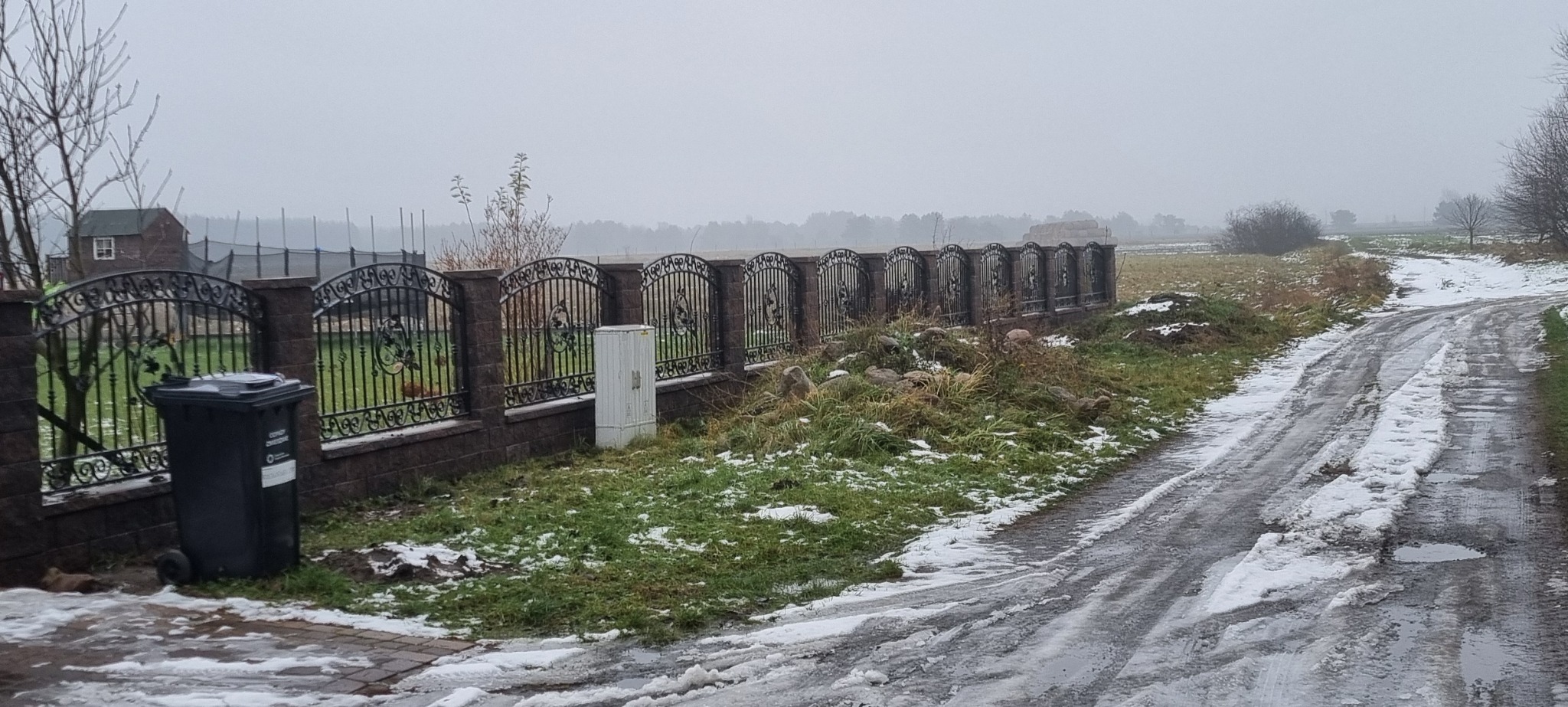 Kute ogrodzenie z dekoracyjnymi elementami, osadzone na niskim murku z cegły. Obok stoi czarny kosz na śmieci, a w tle widać zalesiony teren i budynek.