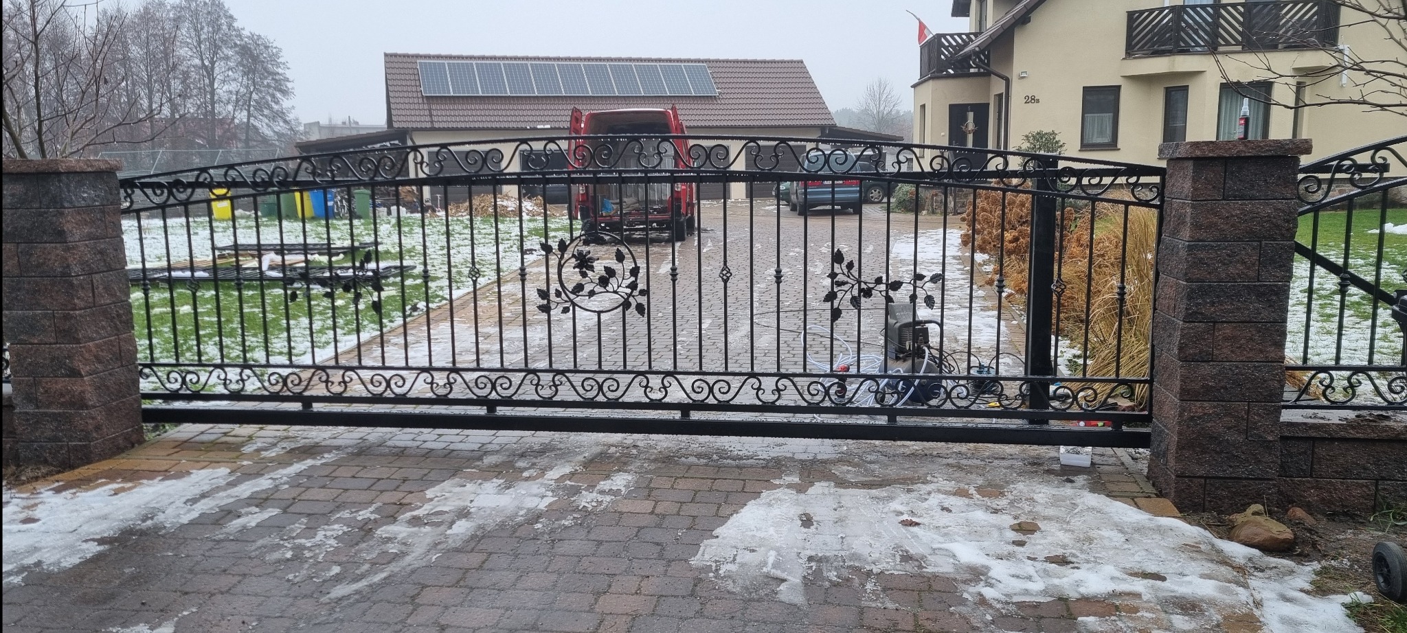 Czarna, kuta brama wjazdowa z dekoracyjnymi elementami roślinnymi, osadzona na murowanych słupkach. W tle dom z panelami słonecznymi i częściowo ośnieżony teren.
