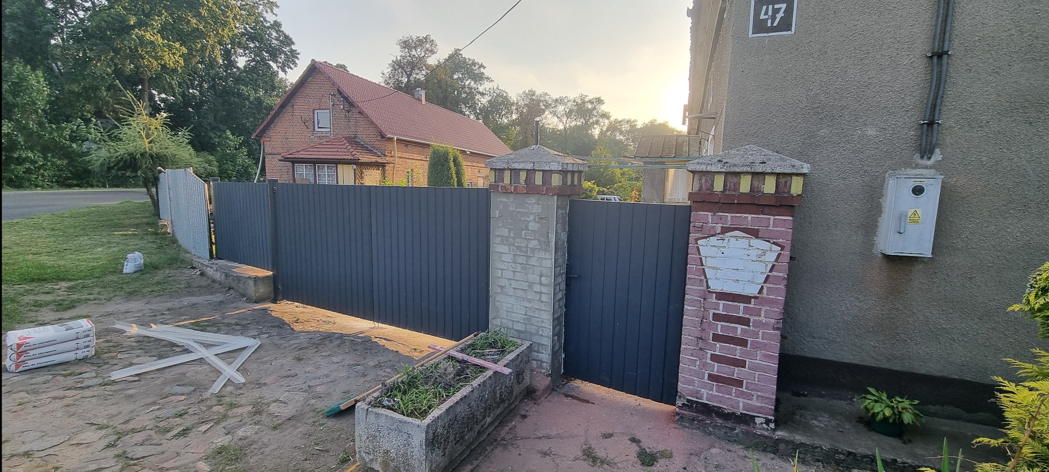 Nowoczesne, grafitowe ogrodzenie panelowe z bramą, osadzone między murowanymi słupkami. W tle dom z czerwonej cegły i zielona roślinność.