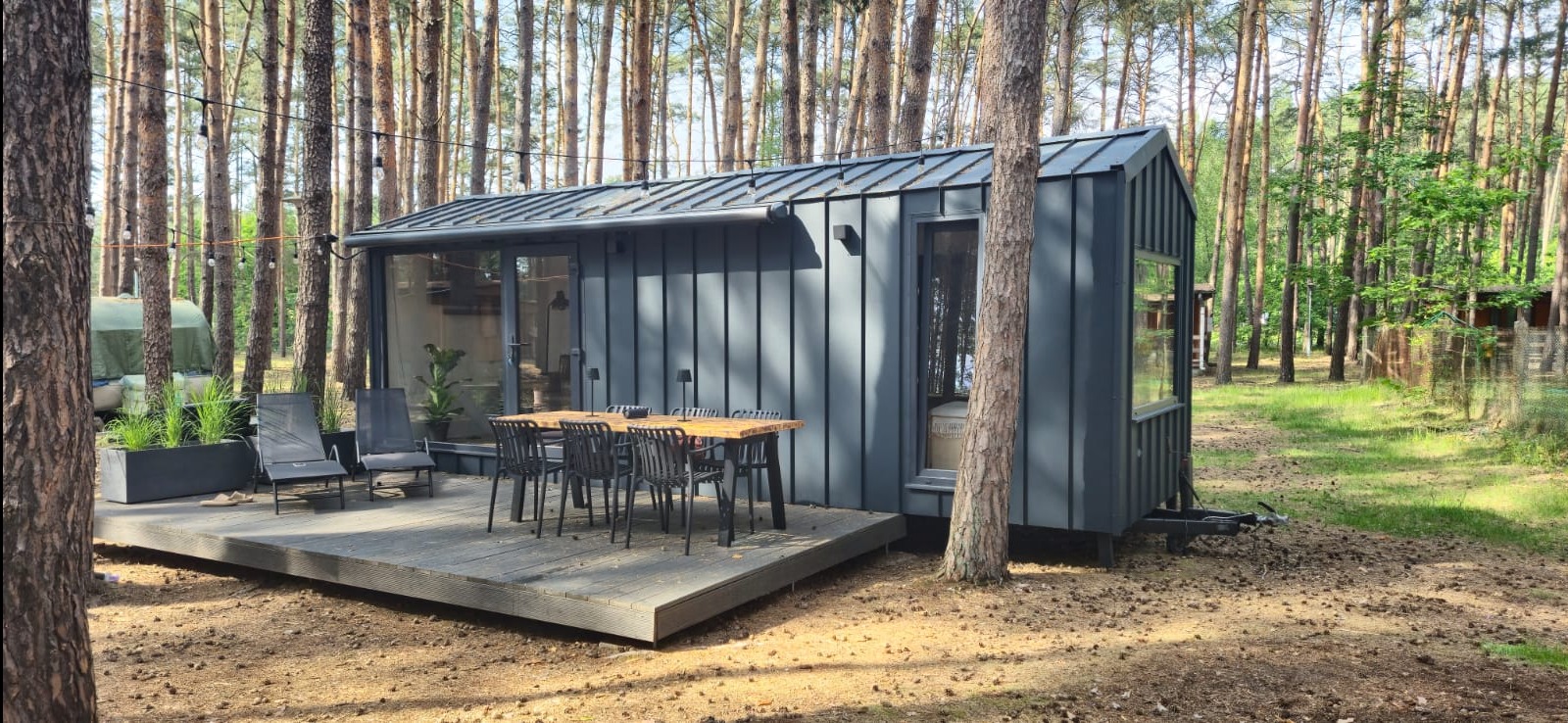 Nowoczesny, szary domek mobilny z metalowym wykończeniem, otoczony sosnowym lasem. Na tarasie drewniany stół z krzesłami i rośliny w donicach. Oświetlenie girlandą żarówek.