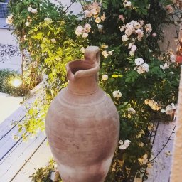 Eco Line Sp. z o.o. - Duży, gliniany dzban ustawiony na drewnianym tarasie, z kwitnącą różą pnącą się po ścianie w tle, oświetloną ciepłym światłem
