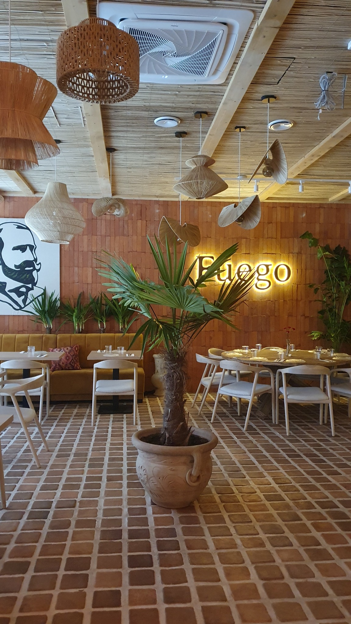 Wnętrze restauracji z ceglaną ścianą, drewnianym sufitem, palmą w ozdobnej donicy na pierwszym planie oraz neonem 'Fuego' w tle.