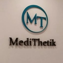 Mangraf Studio - Logo firmy MediThetik z niebieskim okręgiem i inicjałami MT umieszczone nad napisem nazwy firmy, przymocowane do białej ściany.