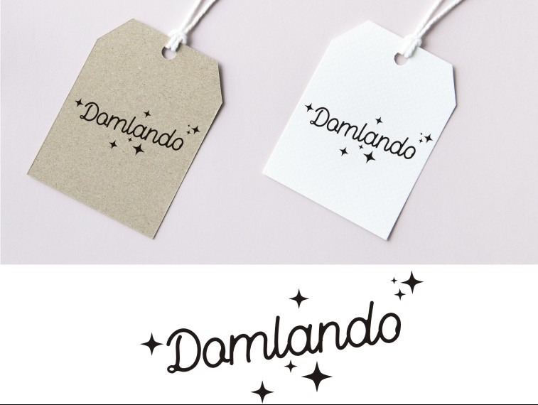 Dwie zawieszki z logo 'Damlando', jedna beżowa, druga biała, leżące na jasnoróżowym tle, poniżej powtórzone logo w czerni.