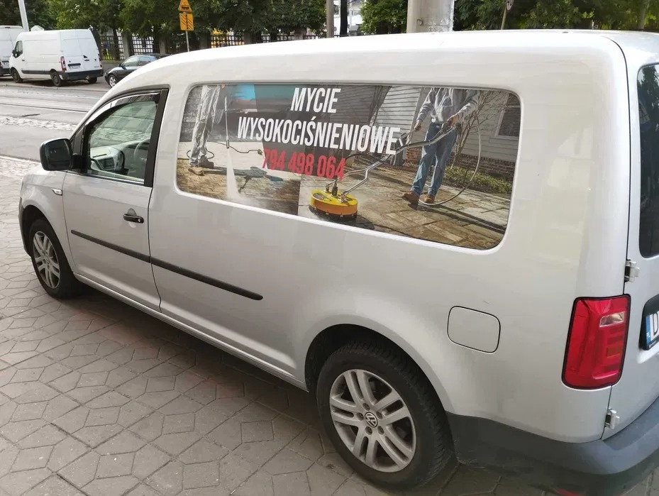 Srebrny samochód dostawczy z reklamą mycia wysokociśnieniowego na bocznych drzwiach, prezentujący osobę czyszczącą powierzchnię za pomocą myjki ciśnieniowej.