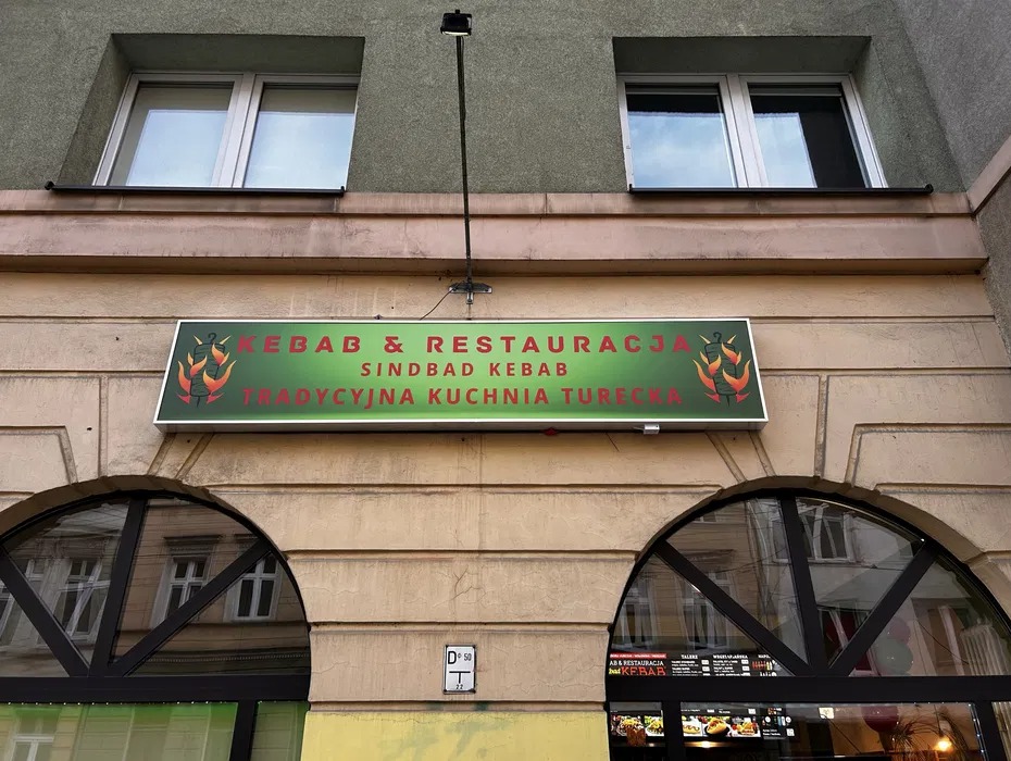 Zewnętrzny szyld restauracji Kebab & Restauracja Sindbad Kebab z motywem płomieni, umieszczony nad łukowym wejściem do lokalu w budynku z jasną elewacją.
