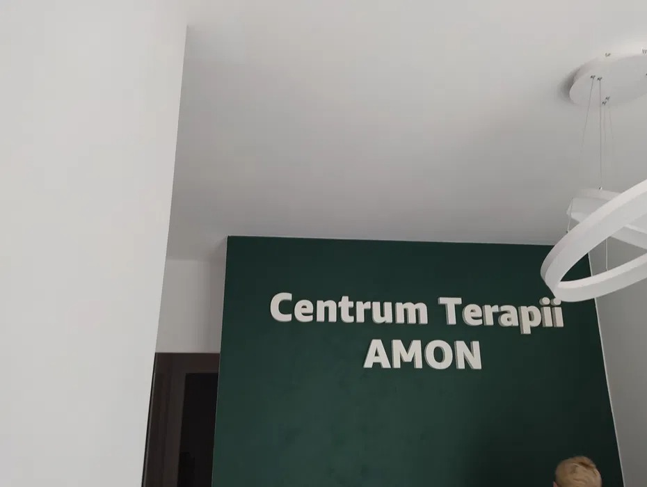 Zielona ściana z napisem 'Centrum Terapii AMON' wykonanym z białych liter 3D, nowoczesna lampa wisząca w tle.