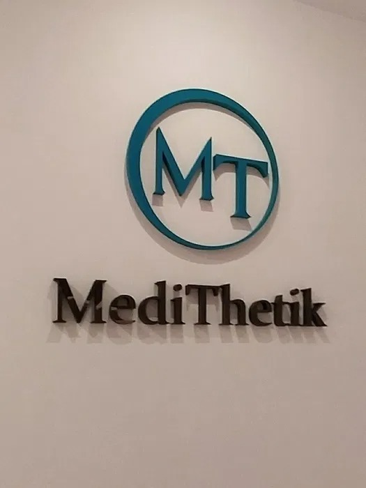 Logo firmy MediThetik z niebieskim okręgiem i inicjałami MT umieszczone nad napisem nazwy firmy, przymocowane do białej ściany.