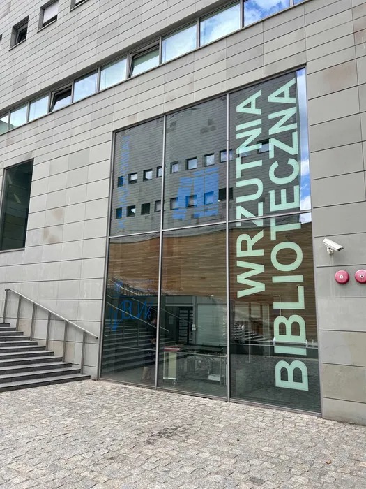 Szklana fasada budynku z napisem 'WRZUTNIA BIBLIOTECZNA' w pionie, widoczne schody wejściowe i szara kostka brukowa na chodniku.