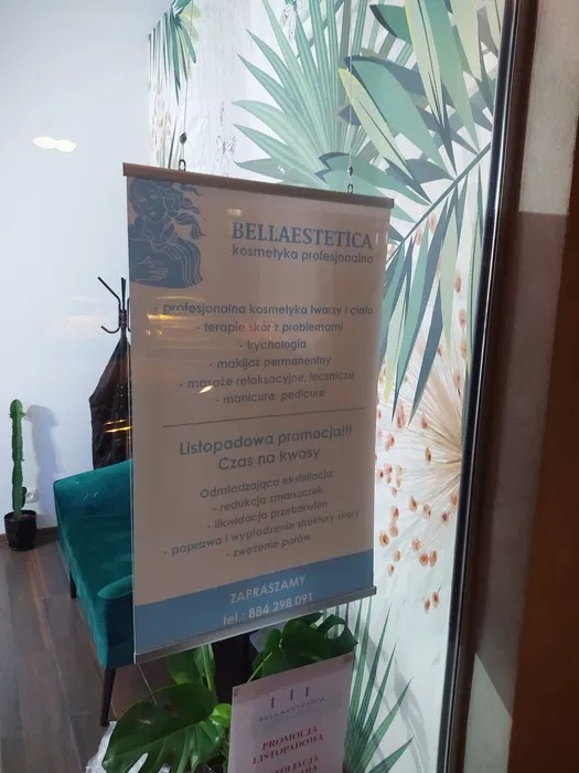 Plakat reklamowy salonu kosmetycznego Bella Estetica z ofertą listopadowej promocji na zabiegi kwasowe, wiszący na tle ściany z motywem tropikalnych liści i zielonego fotela w tle.