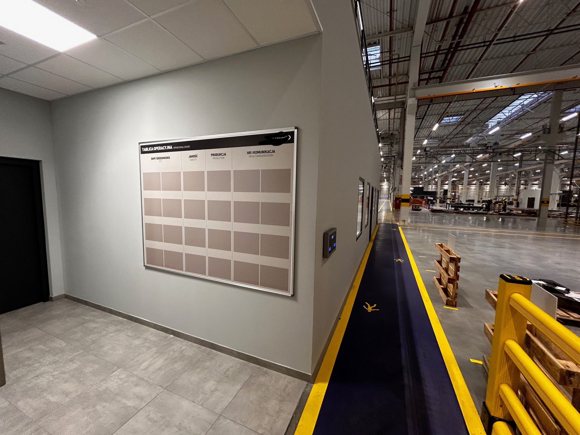 Tablica operacyjna z matrycą pól dla BHP i środowiska, jakości, produkcji oraz HR i komunikacji, umieszczona na ścianie w hali produkcyjnej