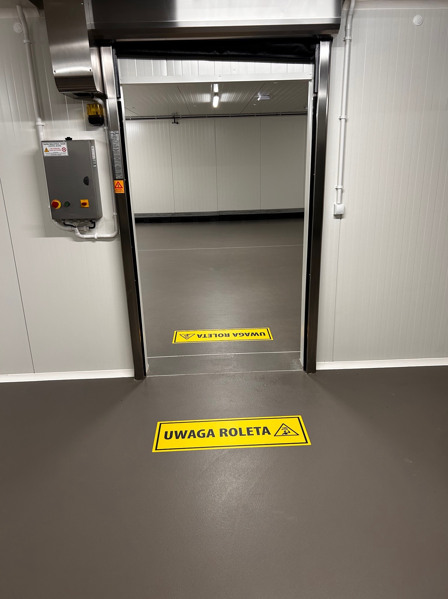 Wnętrze hali produkcyjnej z roletą przemysłową, widoczne ostrzegawcze naklejki podłogowe 'UWAGA ROLETA' w kolorze żółtym z symbolem ostrzegawczym.
