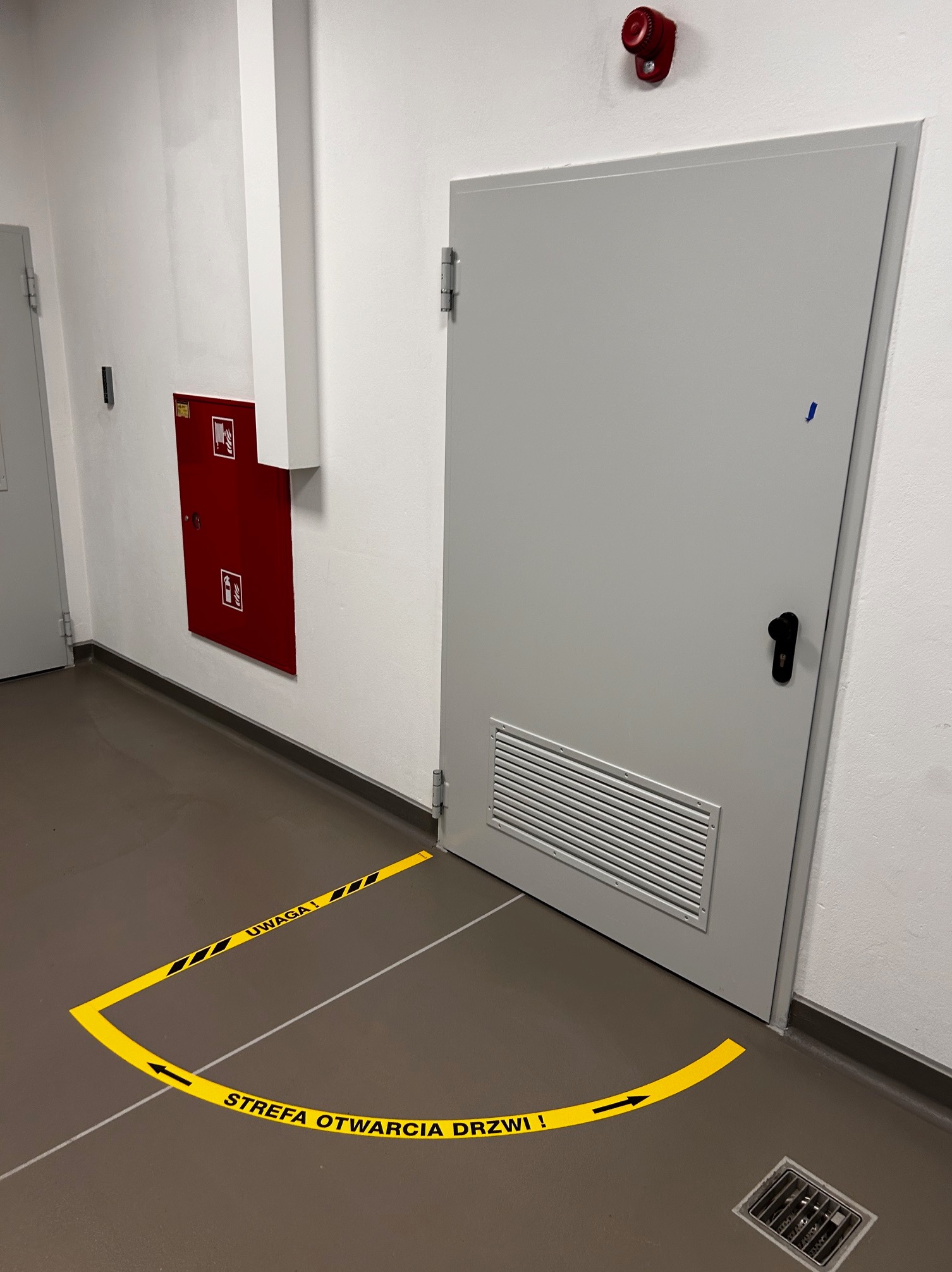 Oznakowanie strefy otwarcia drzwi za pomocą żółtej taśmy ostrzegawczej na podłodze w korytarzu, widoczna metalowa skrzynka hydrantowa na ścianie.