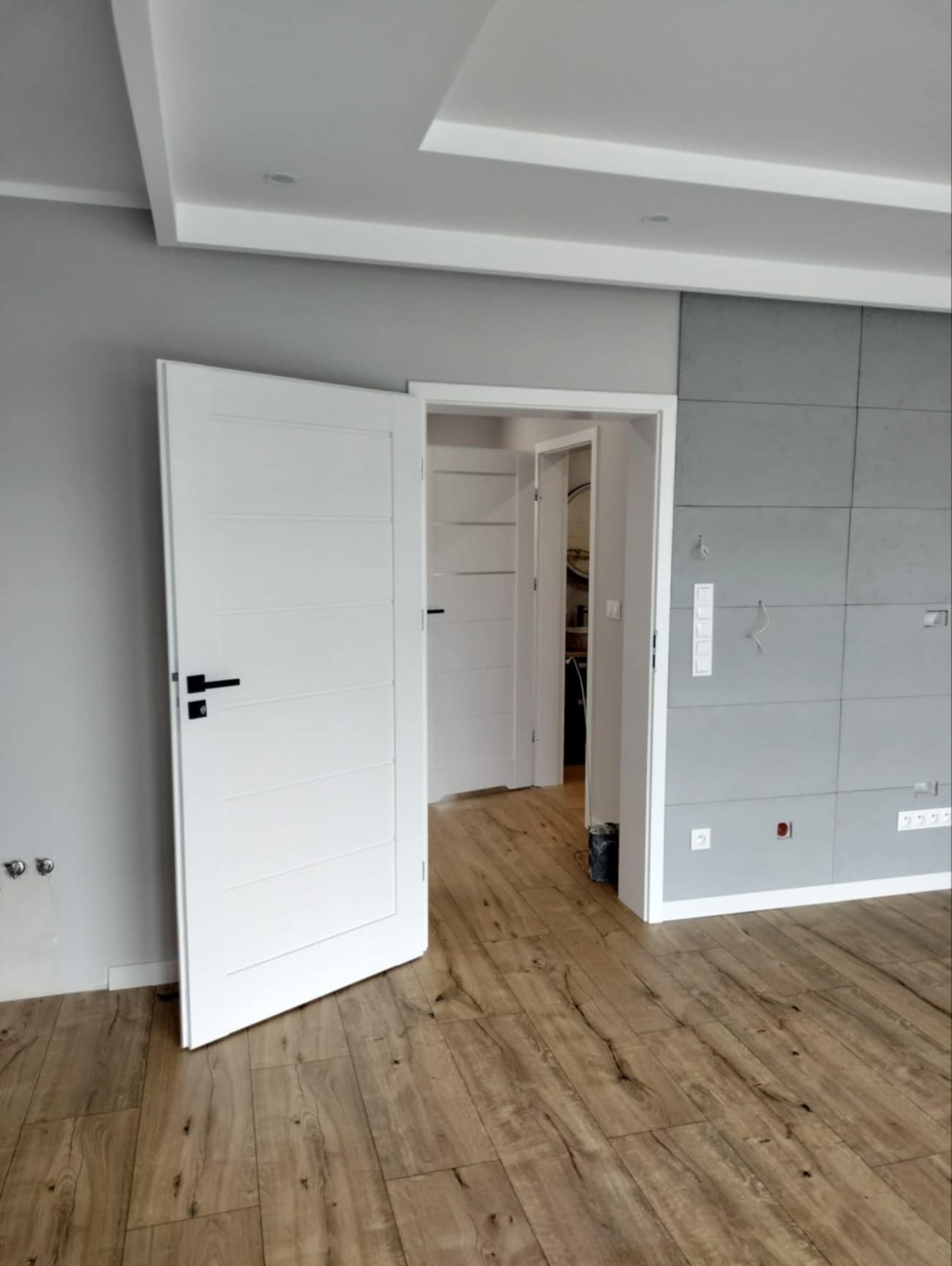 Nowoczesne wnętrze z otwartymi białymi drzwiami z czarną klamką, widoczne panele podłogowe w odcieniach brązu i szara ściana z płytami imitującymi beton.