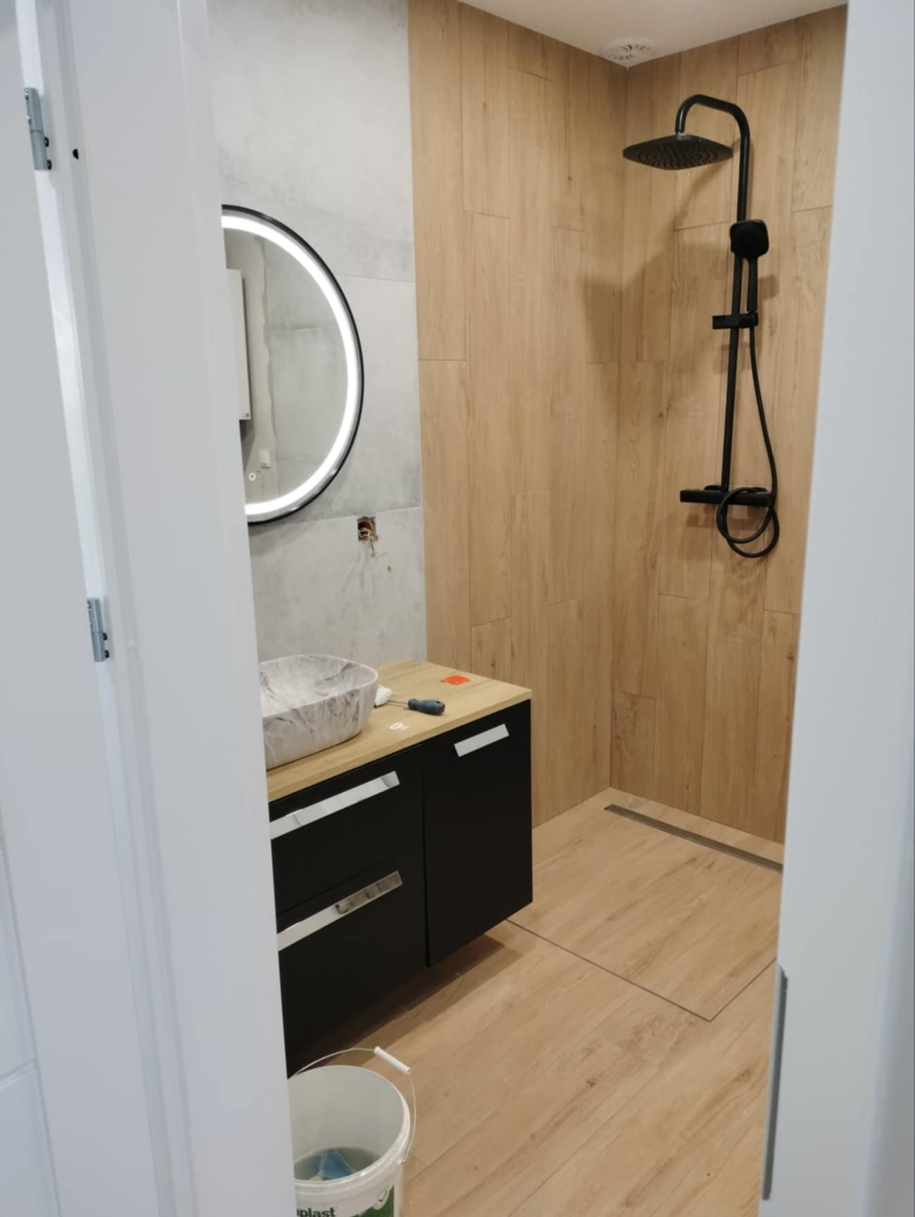 Nowoczesna łazienka z kabiną prysznicową typu walk-in wykończona panelami imitującymi drewno, czarna armatura, umywalka w odcieniach szarości na szafce z czarnymi frontami i drewnianym blatem...