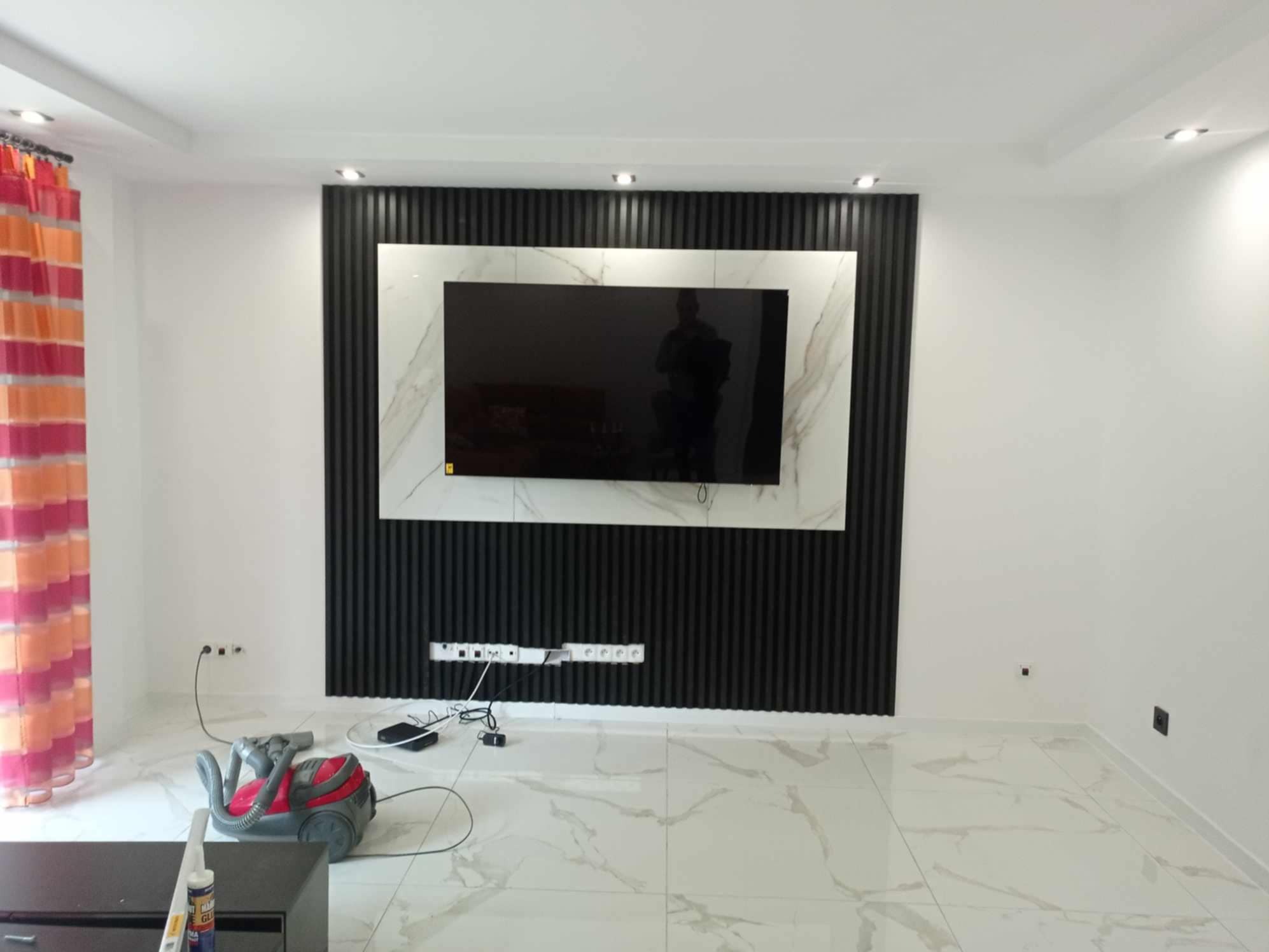 Nowoczesny salon po remoncie: czarny panel lamelowy za telewizorem z marmurową ramą, białe ściany, marmurowe płytki podłogowe, widoczny odkurzacz i listwa z gniazdkami elektrycznymi.
