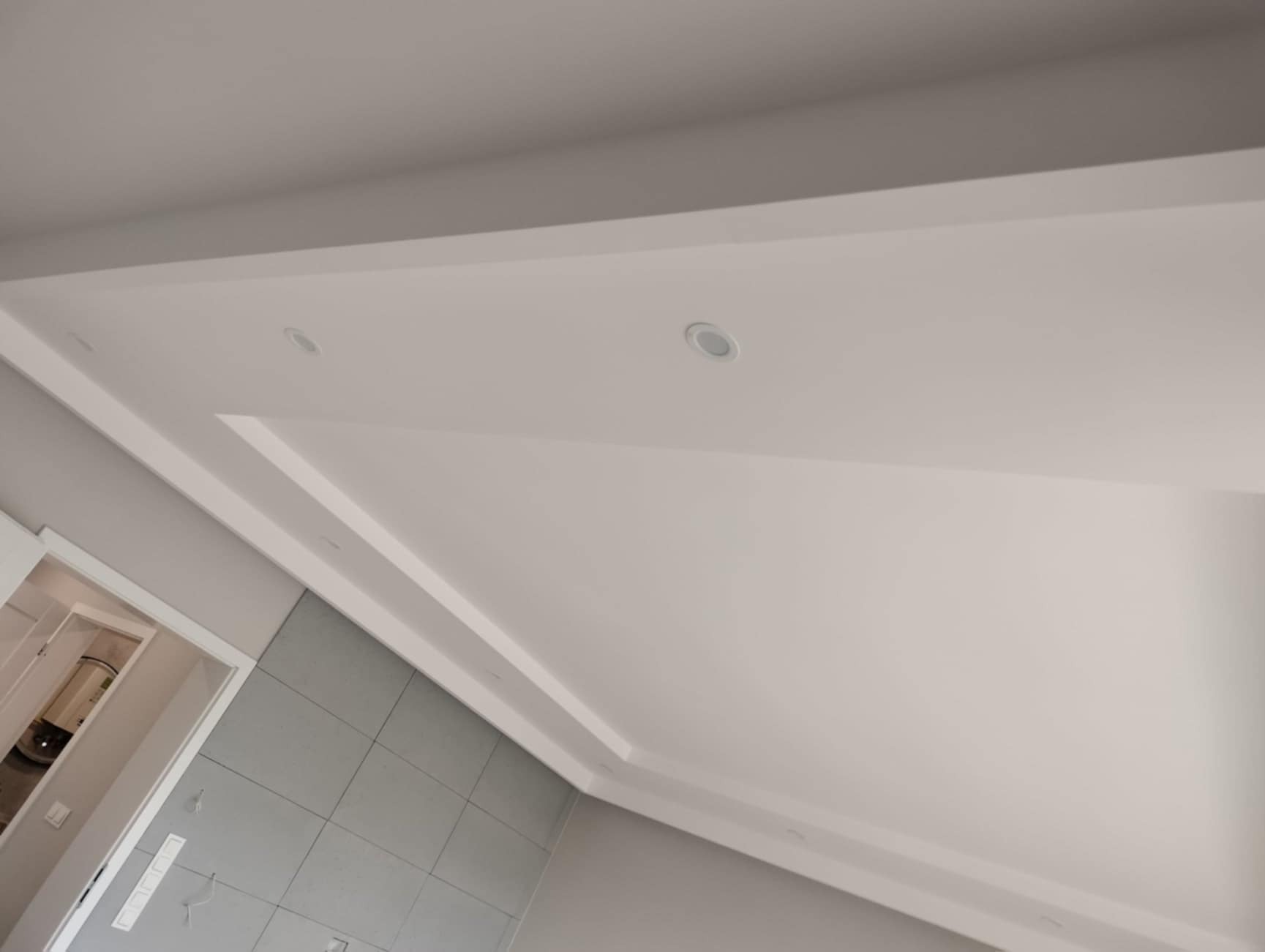 Biały sufit podwieszany z dwoma wpuszczonymi lampami LED i ozdobnym obramowaniem, widok z dołu, szare płytki na ścianie w tle.