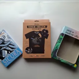 Trzy pudełka produktów: niebieskie z grafiką pary i napisem SICURA, kartonowe z koszulką męską z motywem 'Ogniem i Mieczem' oraz zielone z napisem aura naturals, na białym tle.