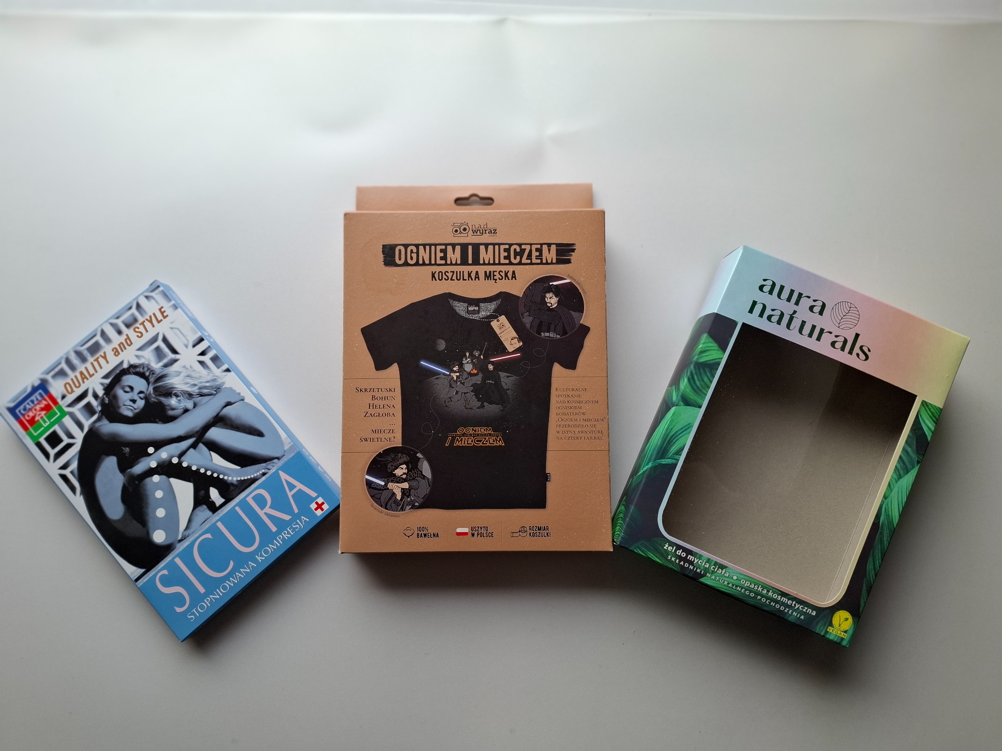 Trzy pudełka produktów: niebieskie z grafiką pary i napisem SICURA, kartonowe z koszulką męską z motywem 'Ogniem i Mieczem' oraz zielone z napisem aura naturals, na białym tle.