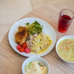 Bistro DoWoli - Kompletny posiłek: kotlet w panierce z brokułami, kaszą gryczaną polaną sosem, pomidorkami cherry, zupą krem i surówką z kapusty, podany na jasnym drewnianym stole.