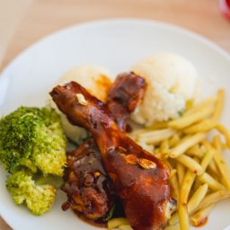 Bistro DoWoli - Porcja obiadowa na białym talerzu: dwa udka z kurczaka w sosie BBQ, kulki ziemniaczane, gotowany brokuł i fasolka szparagowa. Widoczny fragment stołu z jasnego drewna.