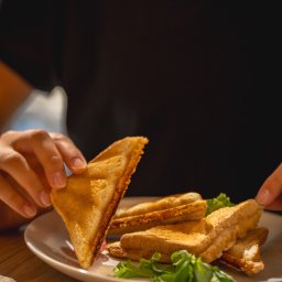 Bistro DoWoli - Osoba trzyma trójkątną kanapkę z tostowego chleba z widocznym nadzieniem, obok talerz z innymi kanapkami i zielonymi listkami sałaty.