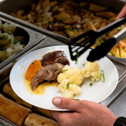 Bistro DoWoli - Nakładanie porcji obiadowej na talerz z bemaru: mięso w sosie, gotowany kalafior i pieczona marchewka, widoczne szczypce i inne potrawy w tle.