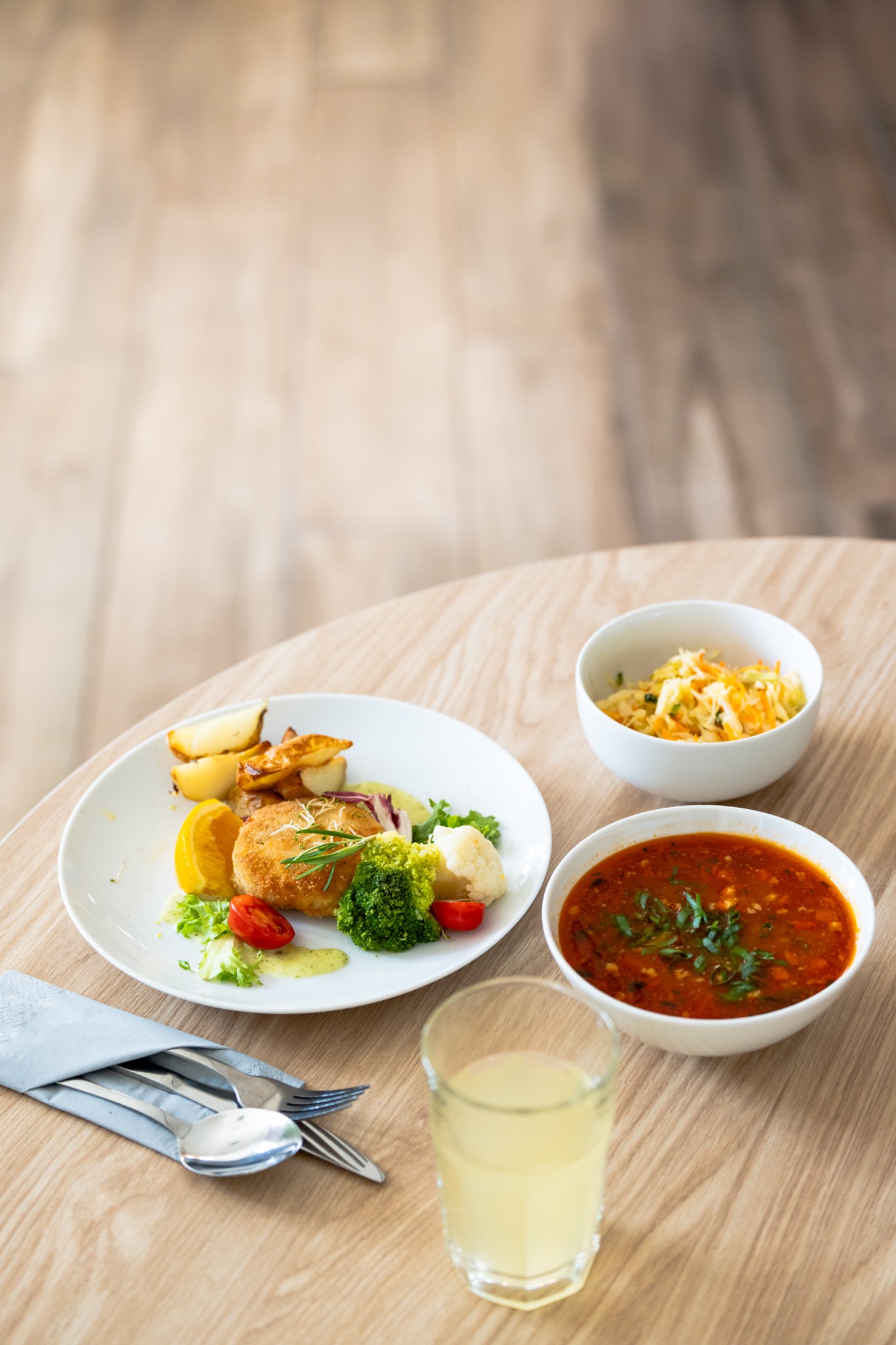 Zestaw obiadowy w Warszawie: kotlet panierowany z ziemniakami, brokułami i sosem, zupa pomidorowa z bazylią oraz surówka z kapusty i marchewki, podane na okrągłym drewnianym stole obok szklanki...