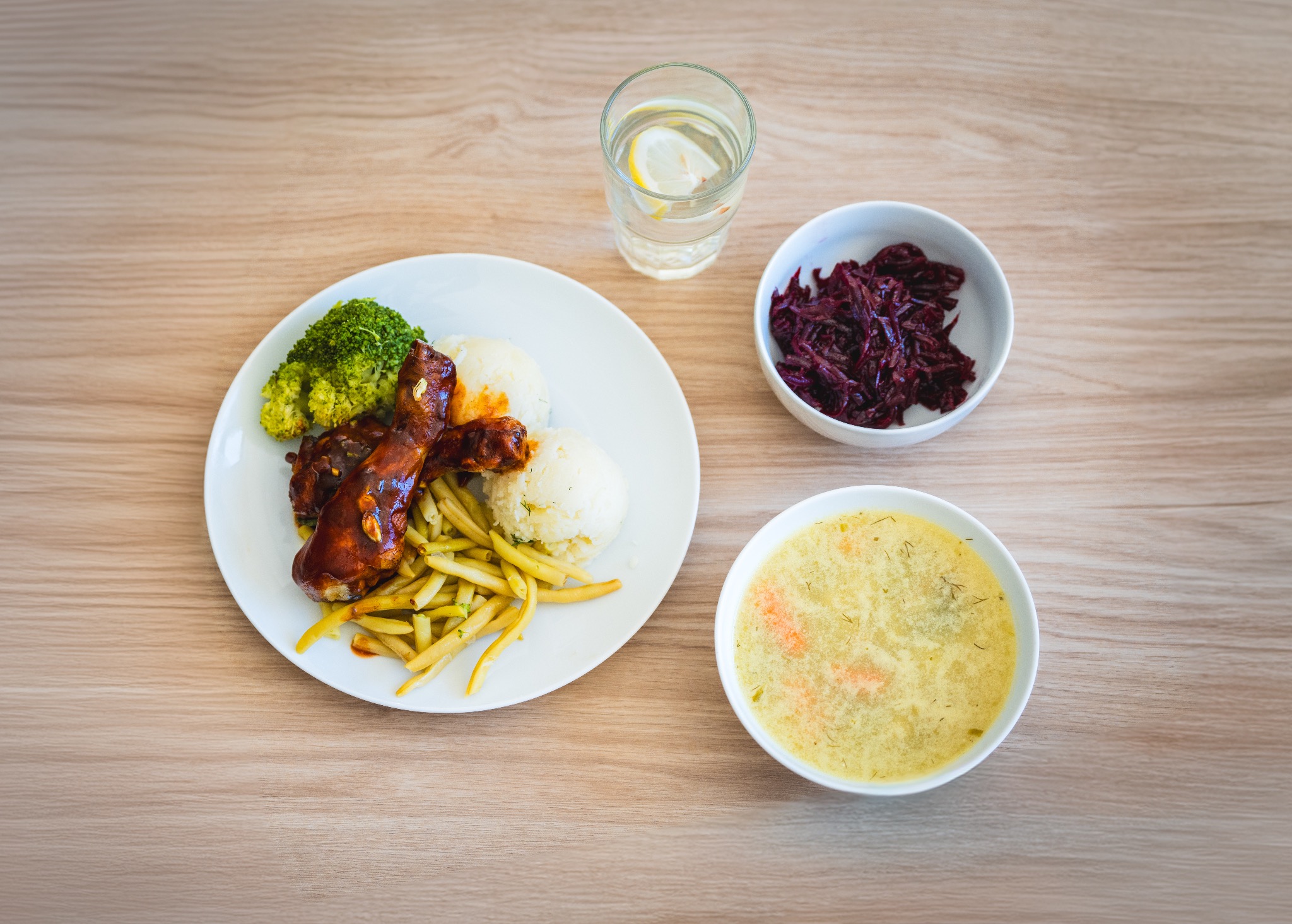 Kompozycja obiadowa: talerz z mięsem w sosie, ziemniakami, brokułami i fasolką szparagową, obok miseczka z czerwoną kapustą, talerz z zupą krem i szklanka wody z cytryną, na jasnym drewnianym stole.