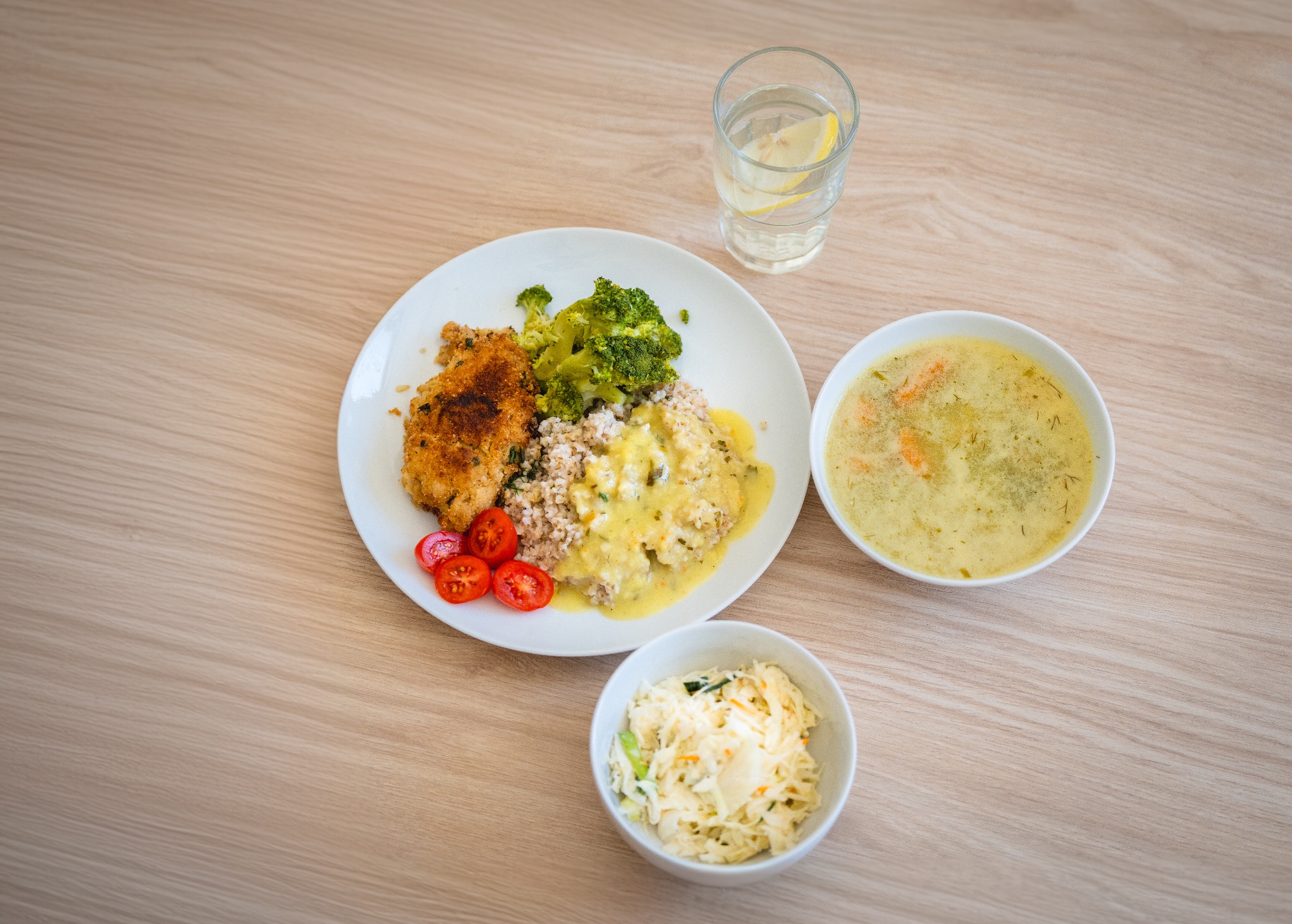 Kompozycja obiadowa: kotlet panierowany, kasza z sosem, brokuły, pomidorki cherry, zupa krem i surówka w białych miseczkach, szklanka wody z cytryną na drewnianym stole.