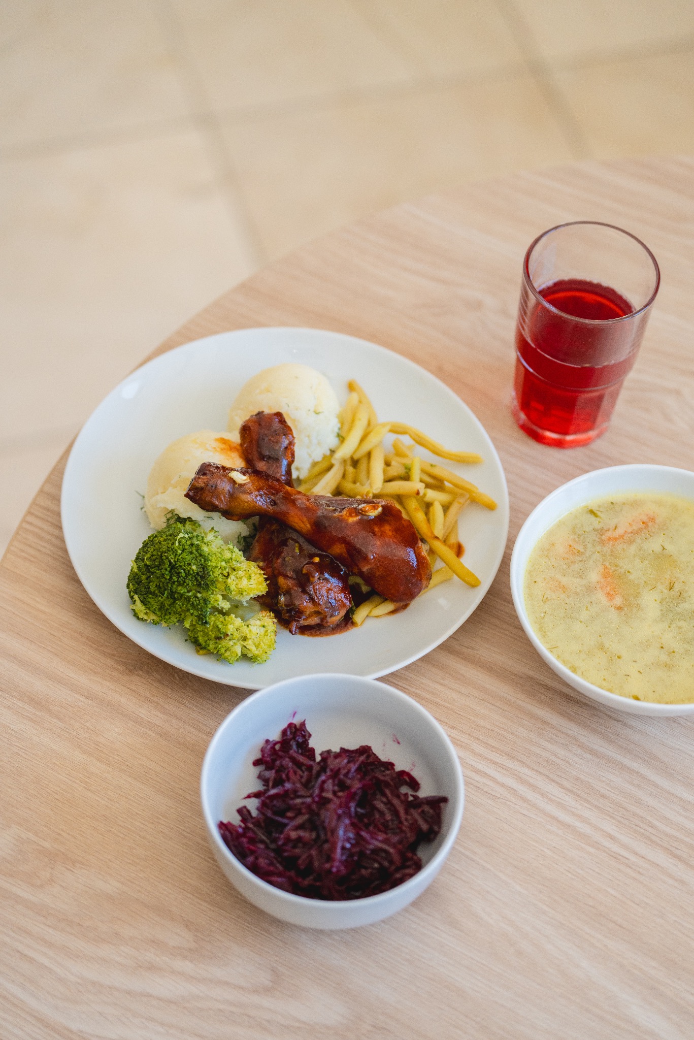 Kompletny obiad na jasnym stole: talerz z dwoma udkami w ciemnym sosie, ziemniakami puree, zieloną fasolką szparagową, różyczką brokuła, czerwona kapusta w miseczce, szklanka czerwonego napoju...