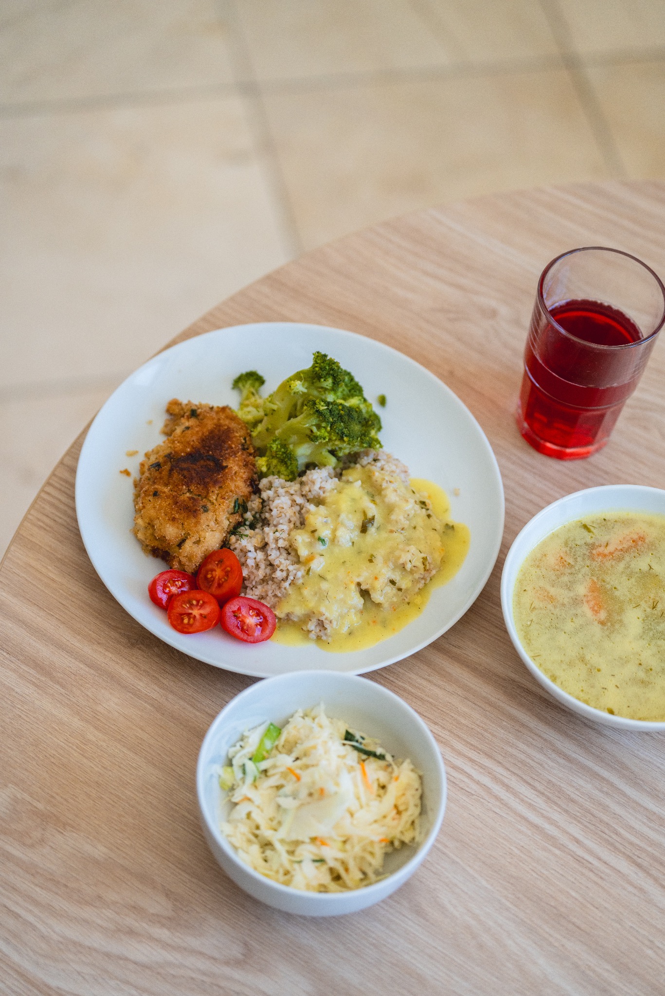 Kompletny posiłek: kotlet w panierce z brokułami, kaszą gryczaną polaną sosem, pomidorkami cherry, zupą krem i surówką z kapusty, podany na jasnym drewnianym stole.