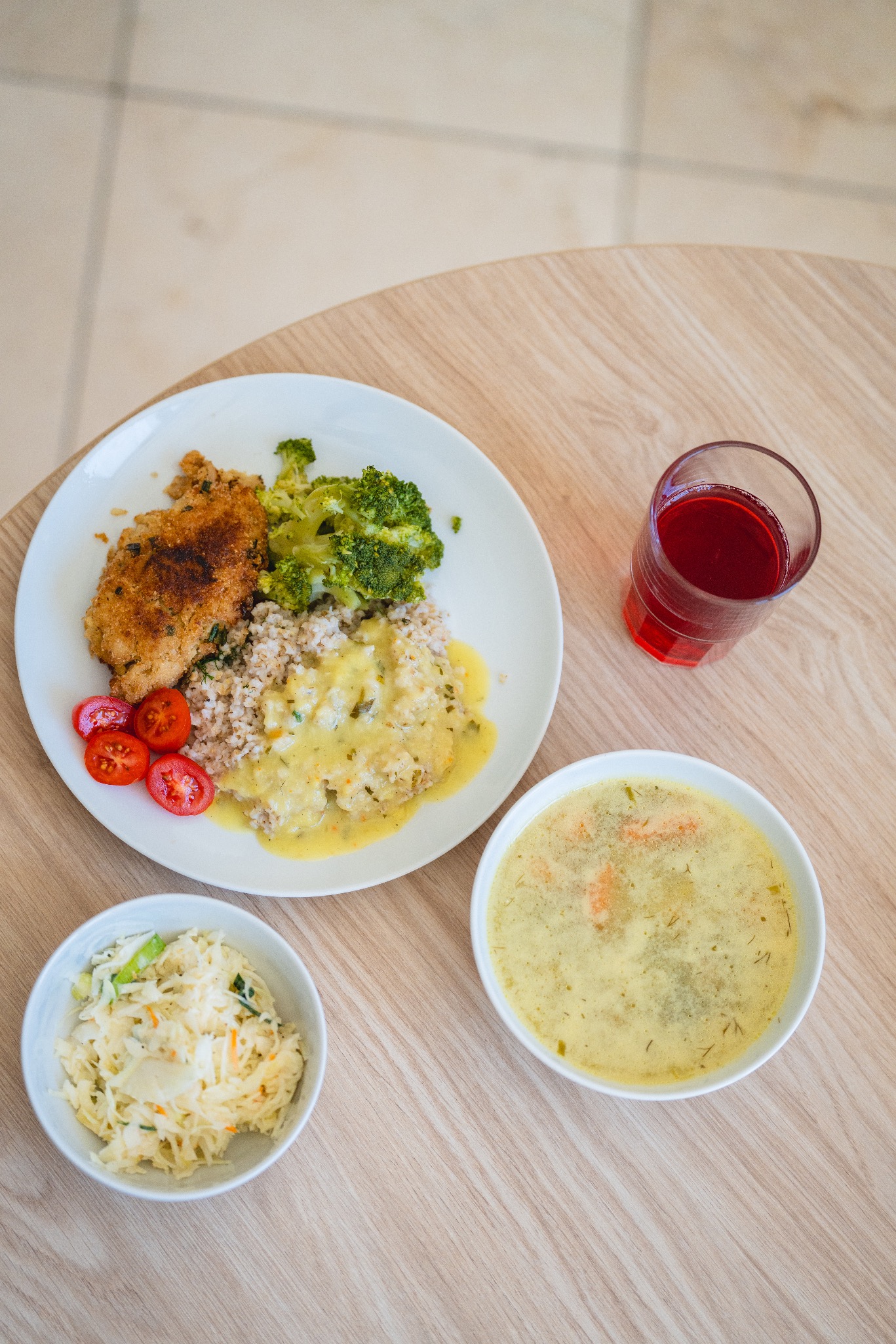 Zestaw obiadowy na jasnym drewnianym stole: kotlet w panierce, kasza z sosem, brokuły, pomidorki koktajlowe, surówka z kapusty i zupa krem, do tego szklanka czerwonego napoju.