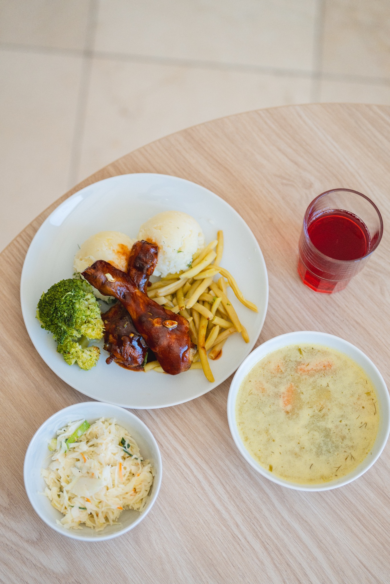 Kompletny posiłek na jasnym drewnianym stole: pieczone udka kurczaka w glazurze, ziemniaki, zielona fasolka szparagowa, brokuły, surówka z kapusty, zupa krem i szklanka czerwonego soku.