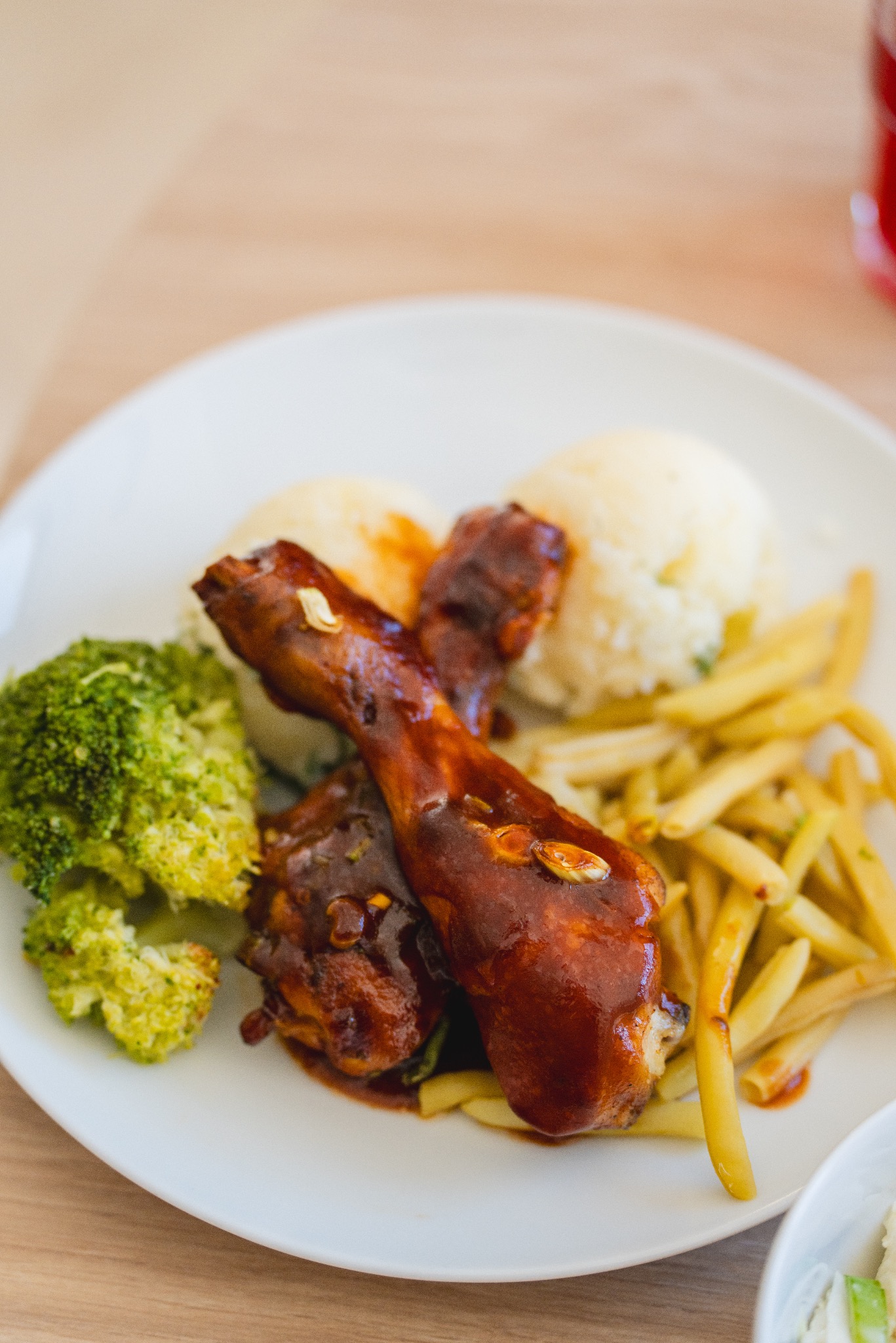 Porcja obiadowa na białym talerzu: dwa udka z kurczaka w sosie BBQ, kulki ziemniaczane, gotowany brokuł i fasolka szparagowa. Widoczny fragment stołu z jasnego drewna.