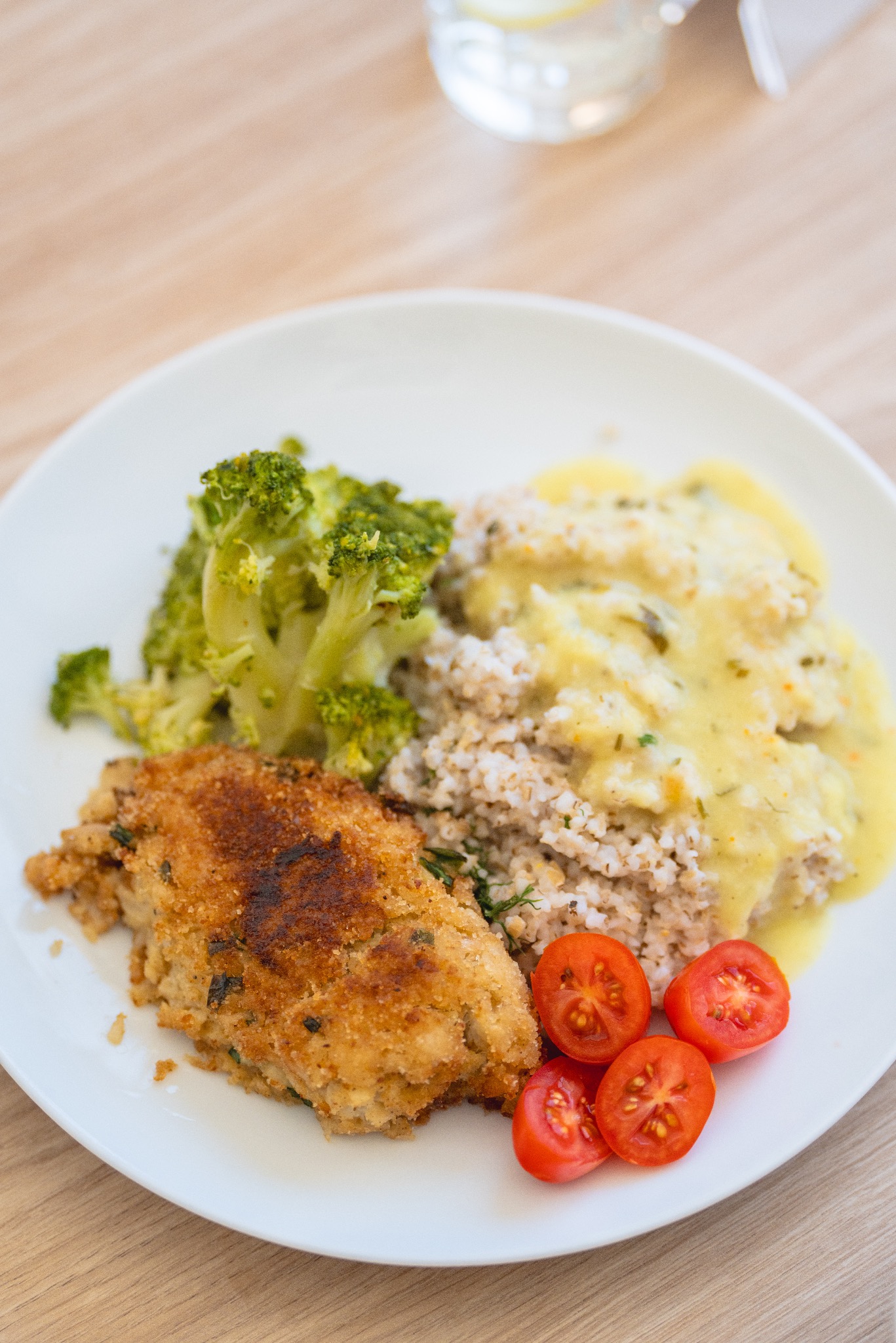 Talerz z kotletem w panierce, brokułami, kaszą gryczaną z sosem i połówkami pomidorków koktajlowych, ułożony na drewnianym stole, w tle szklanka z wodą i cytryną.