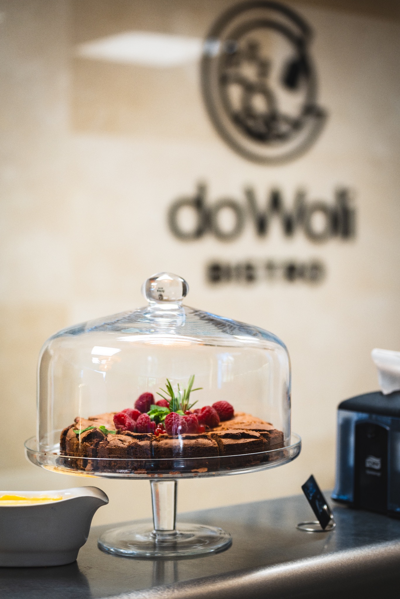 Czekoladowe ciasto na szklanej paterze pod kloszem, udekorowane malinami i gałązką rozmarynu, na tle ściany z logo bistro.