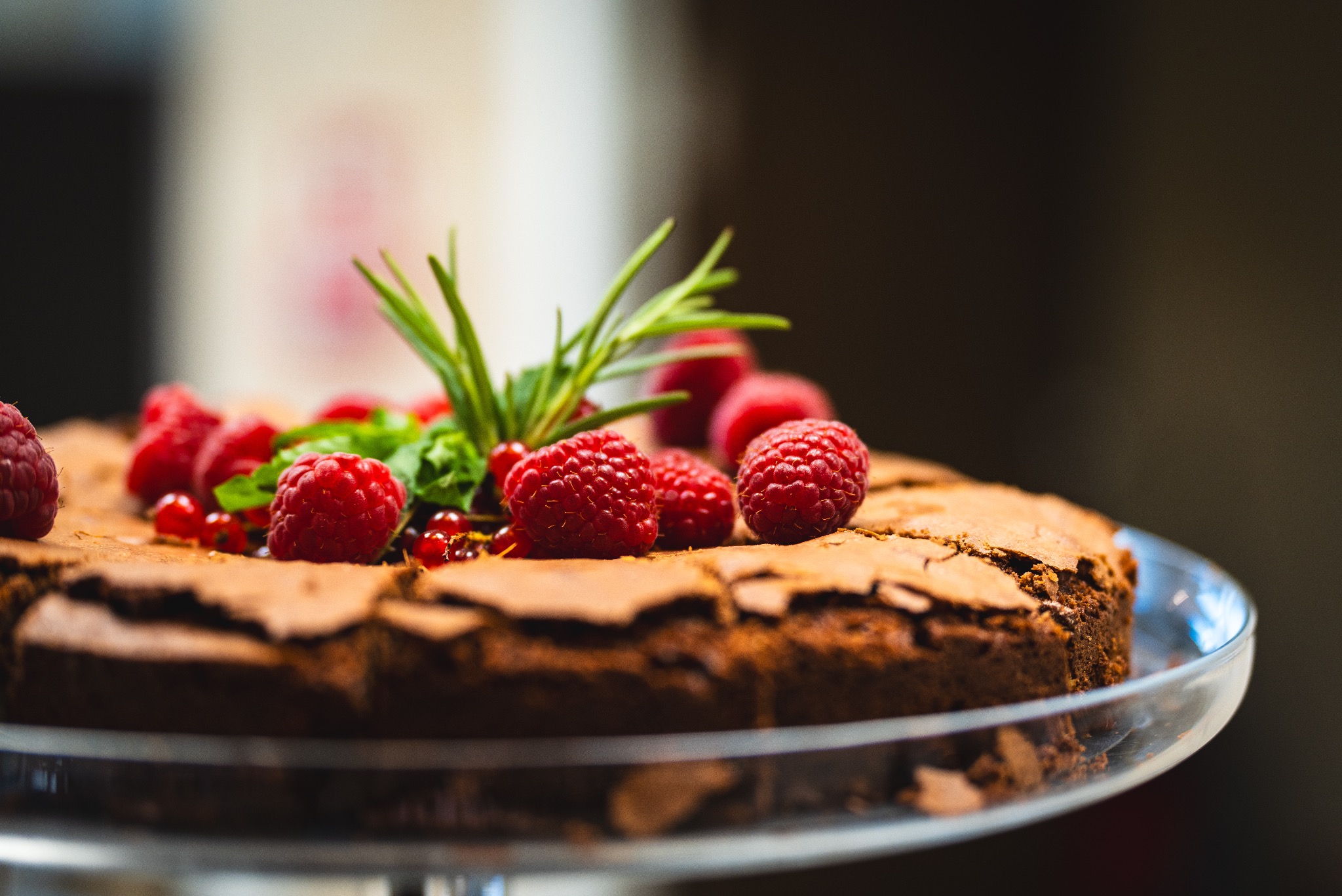 Czekoladowe ciasto brownie udekorowane świeżymi malinami, czerwoną porzeczką i gałązką rozmarynu, podane na szklanym paterze.