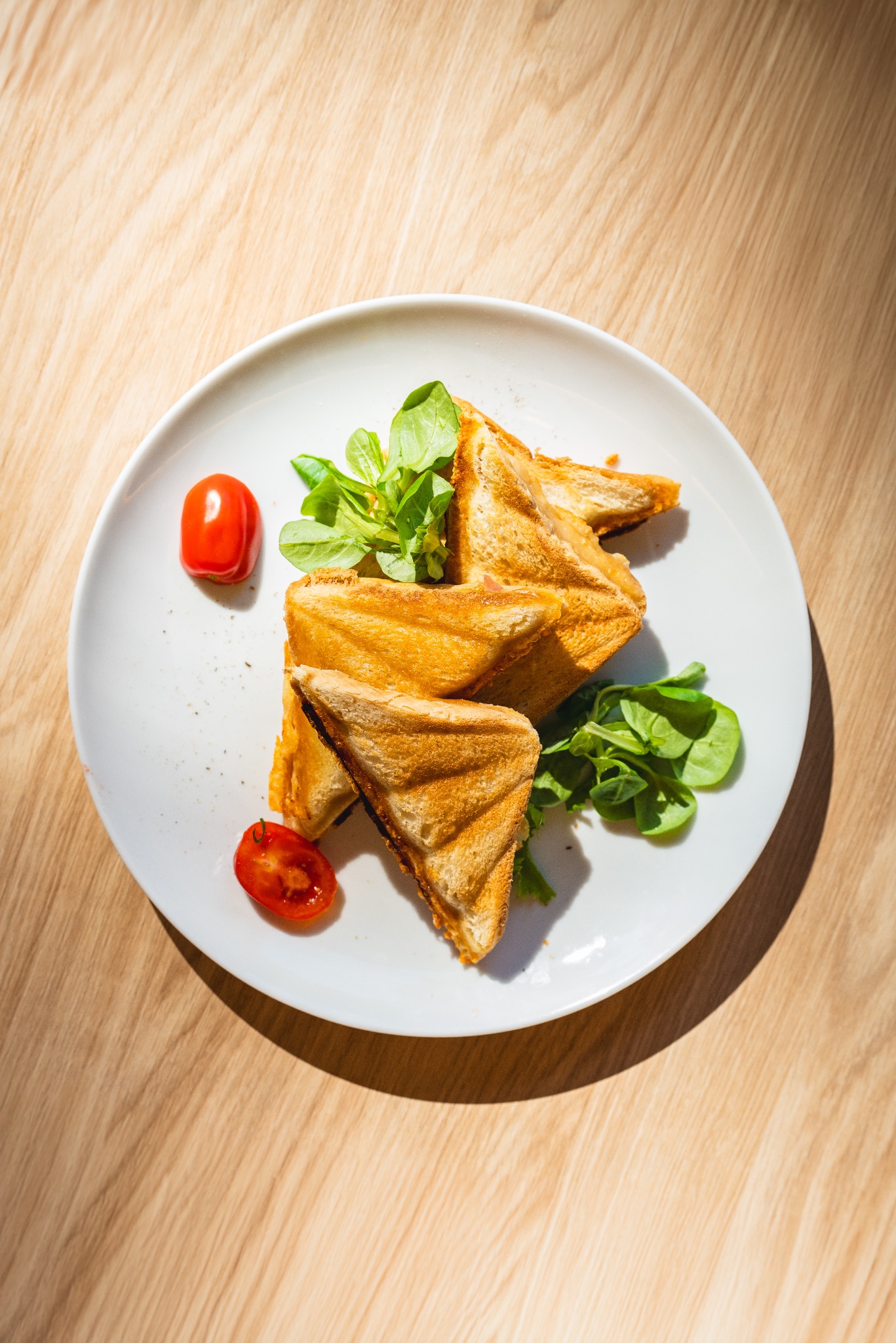 Trójkątne tosty z grilla na białym talerzu, udekorowane świeżą sałatą i połówkami pomidorków cherry, oświetlone jasnym światłem słonecznym na drewnianym stole.